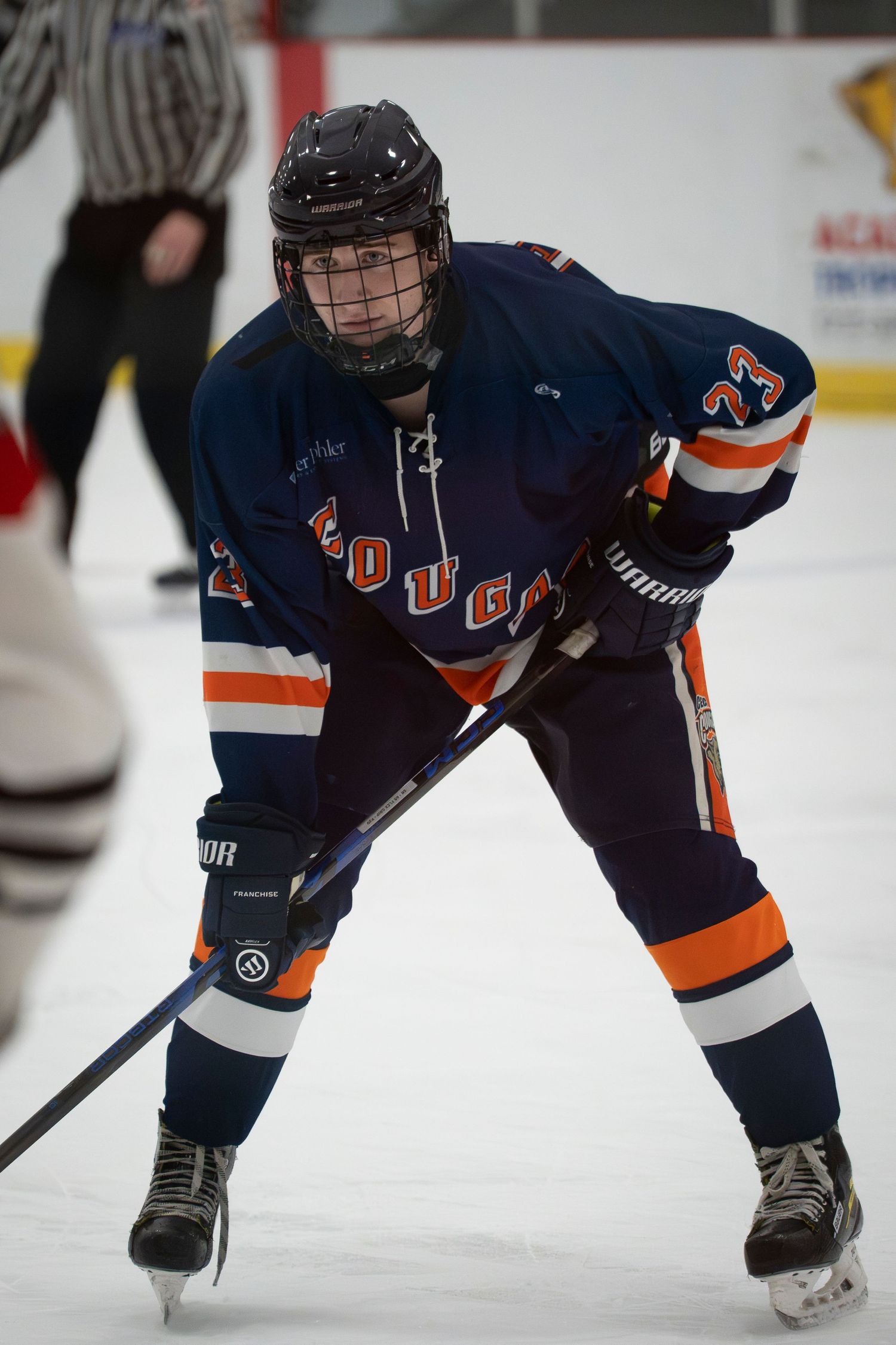 Youth hockey player in navy blue and orange uniform skating on ice during a game.