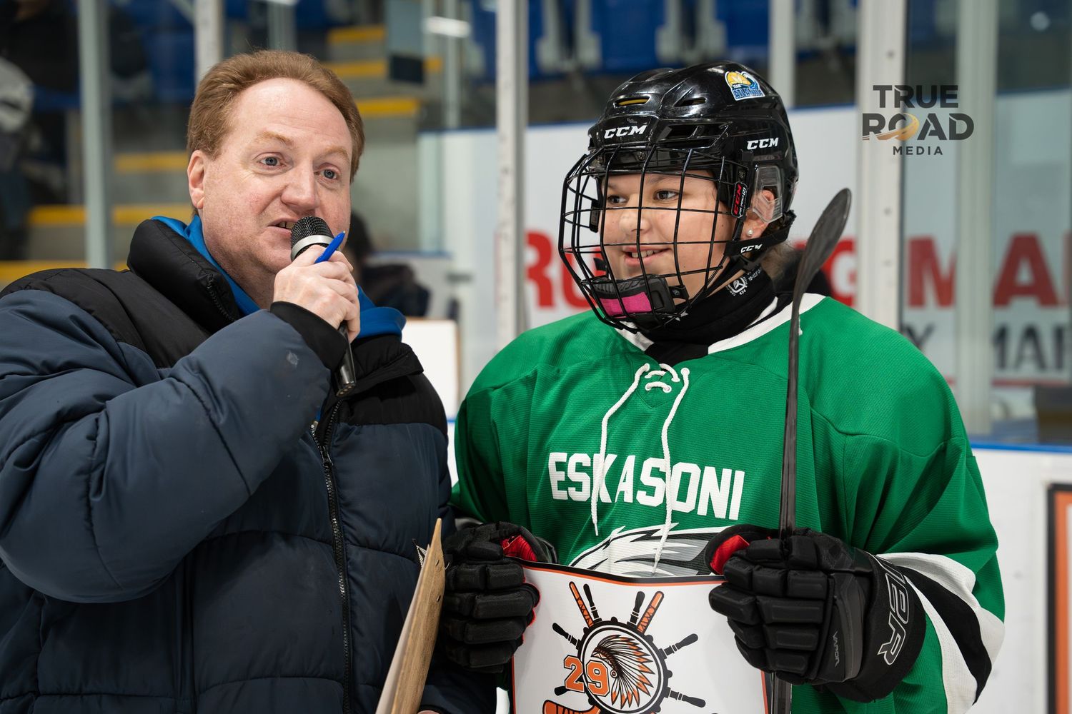Reporter with microphone interviews hockey player wearing green Escanoni jersey at ice rink.