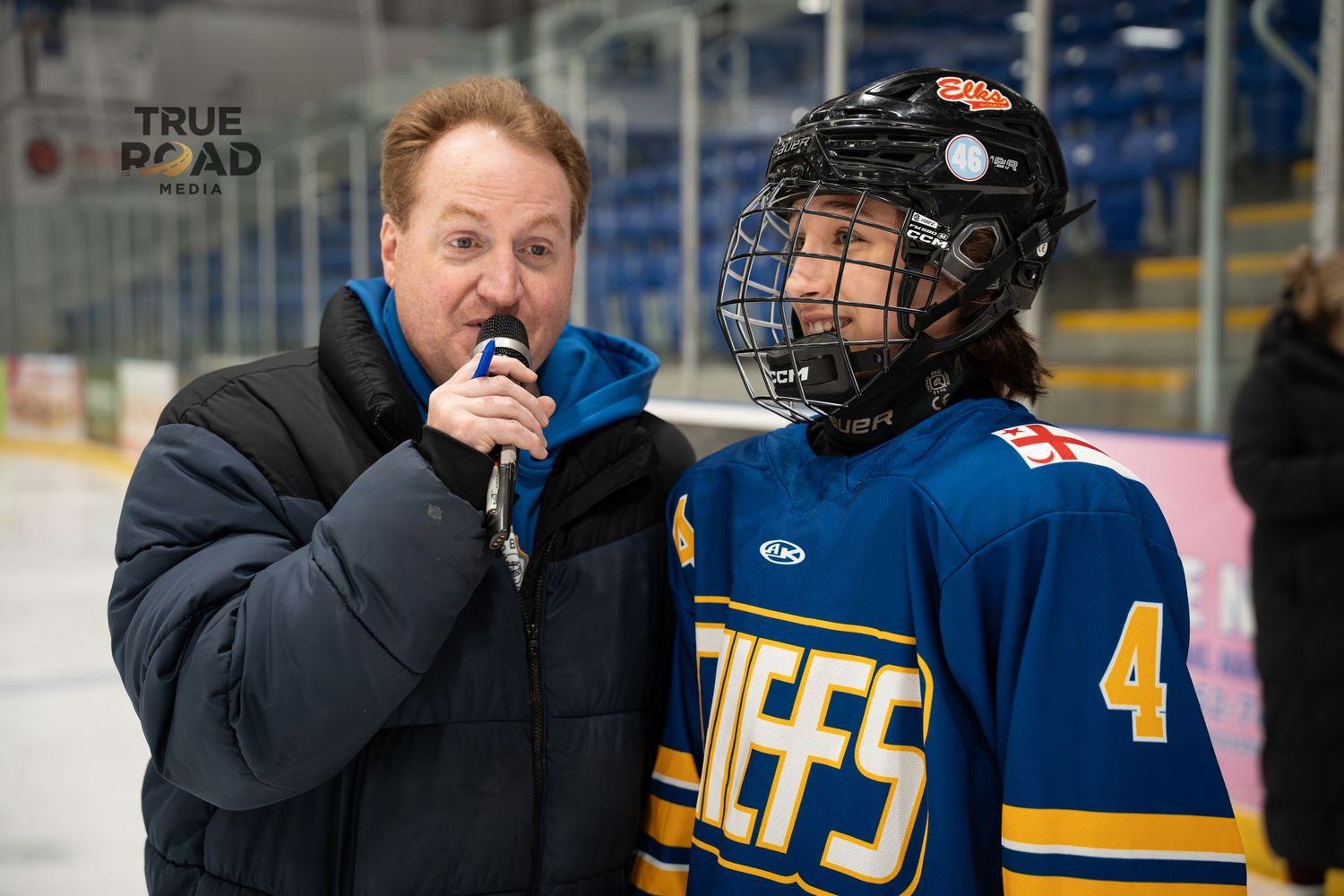 Announcer speaks into microphone next to hockey player during event.