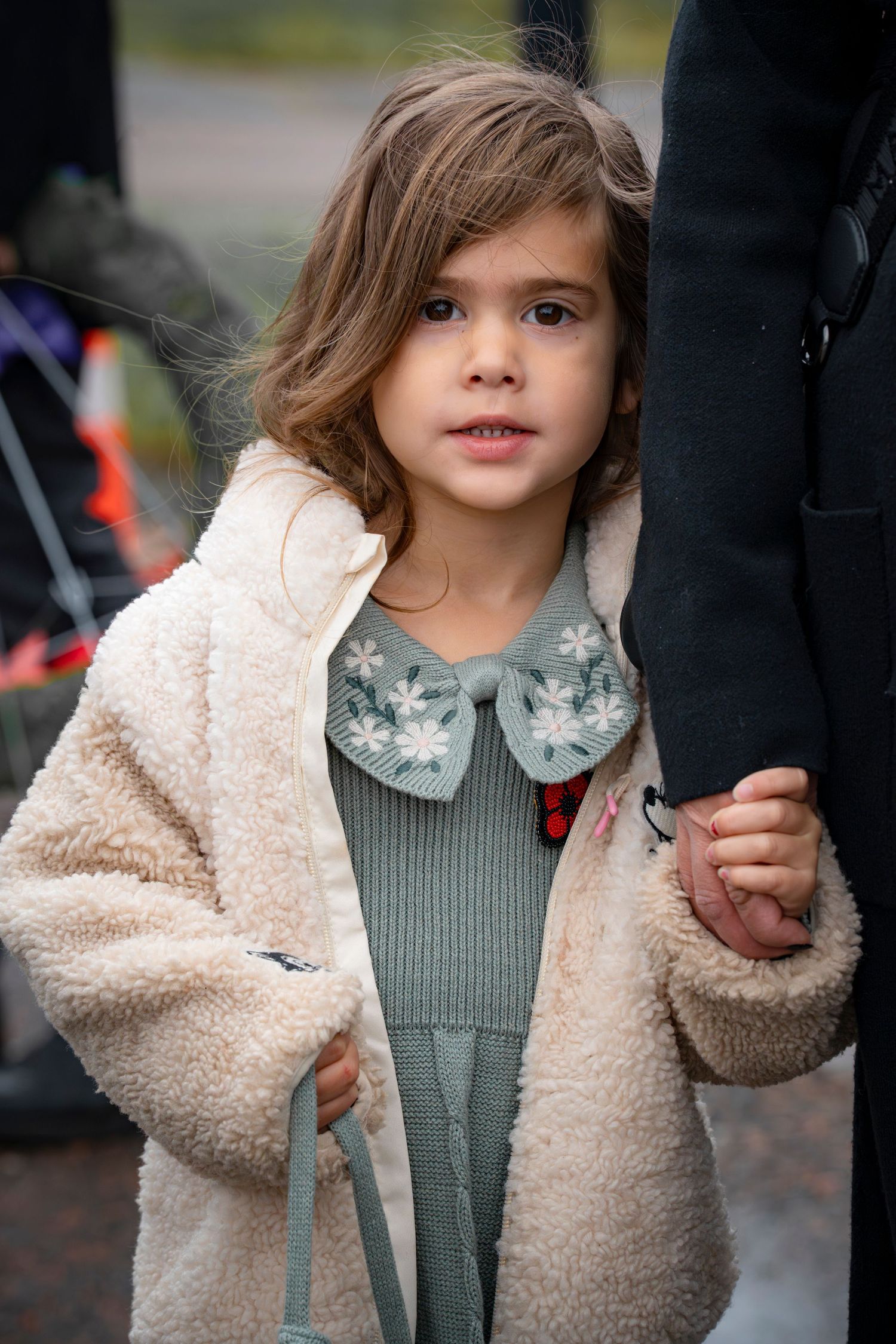 A child in a cozy cream sherpa jacket and green dress holds someone's hand while walking outdoors.