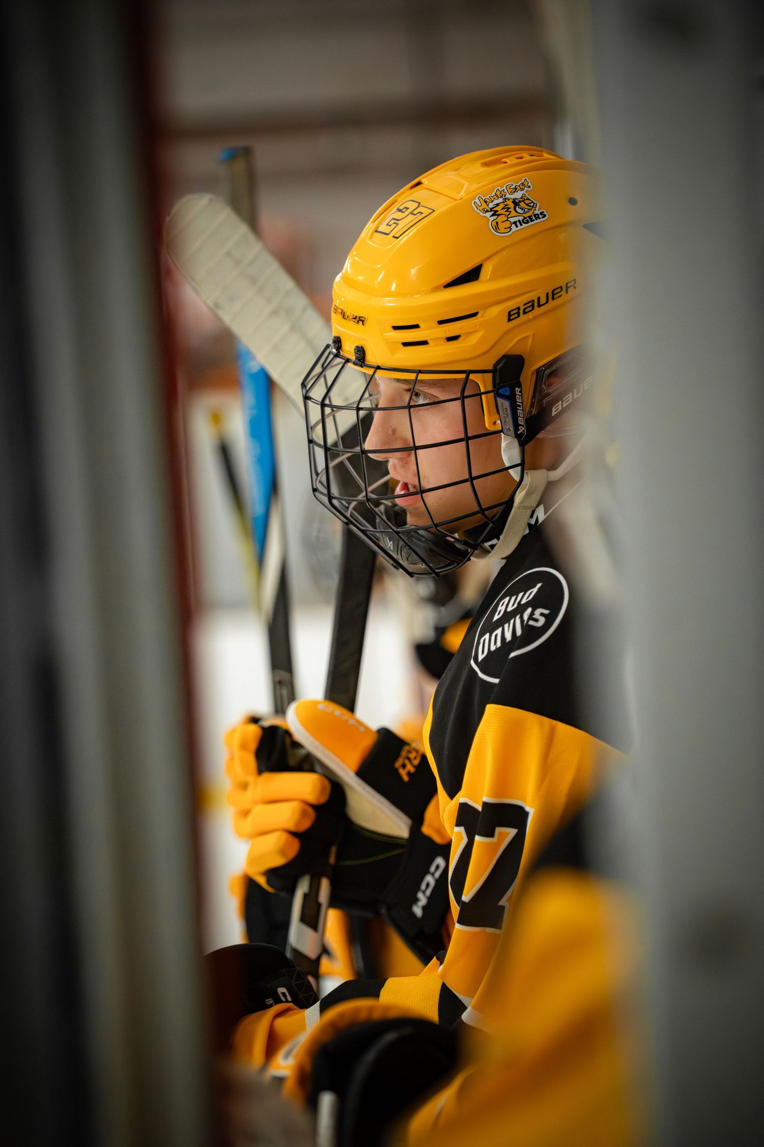Hockey player in yellow jersey and helmet with cage face mask holds stick while sitting on bench.
