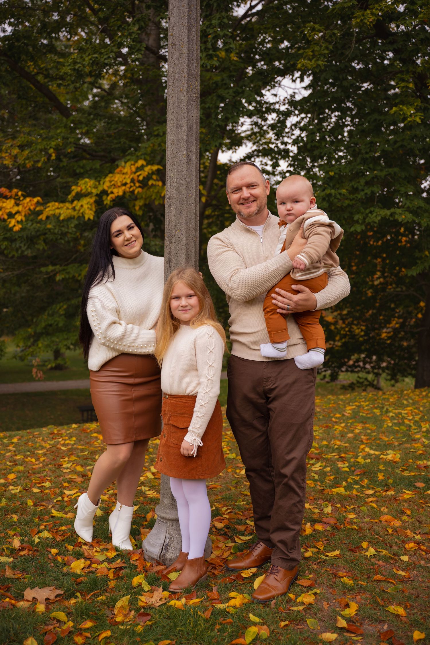 A family in coordinating cream and brown outfits poses together on fallen autumn leaves in an outdoor setting.
