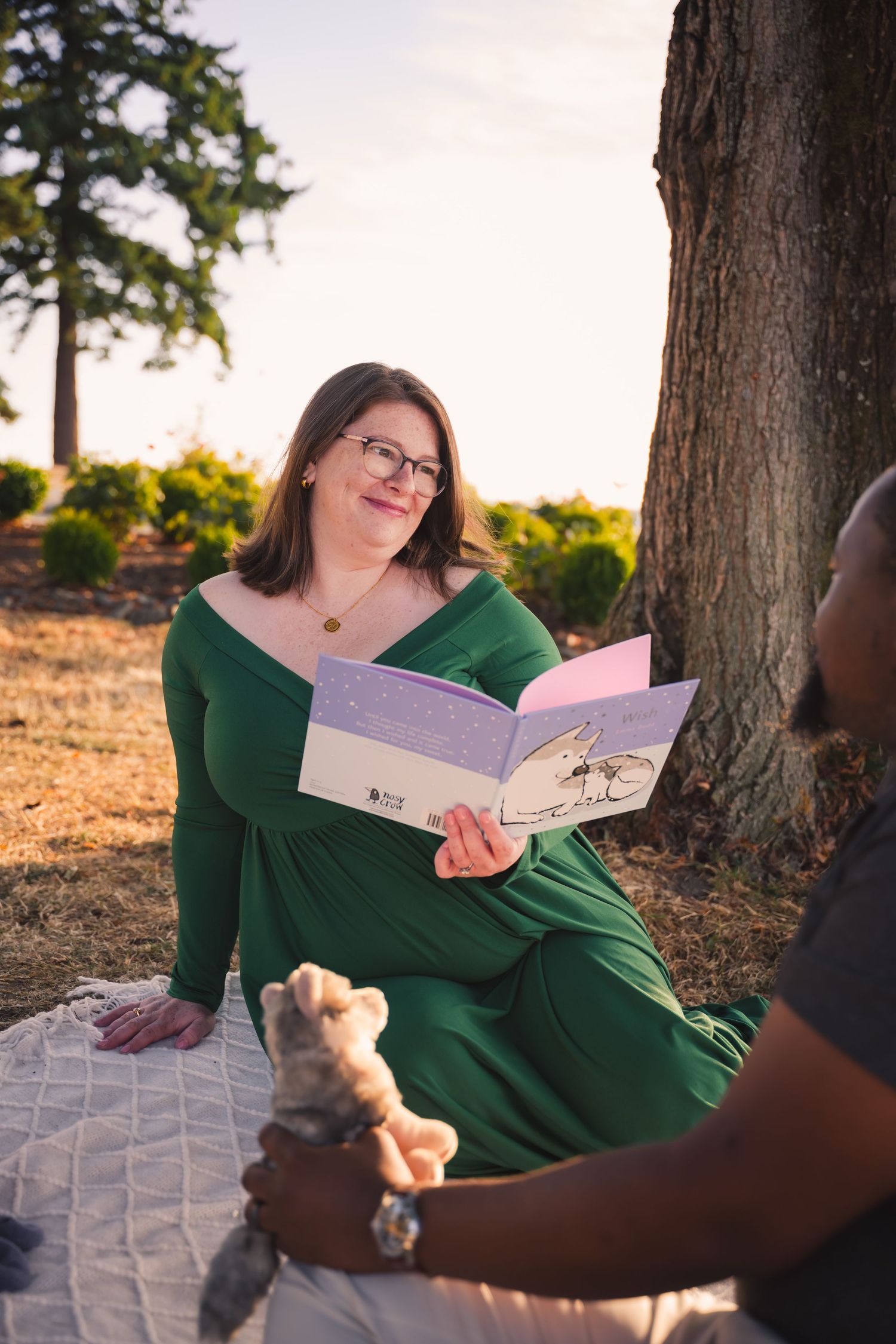 Person in green sweater reading a book while relaxing outdoors on a blanket near trees at sunset.