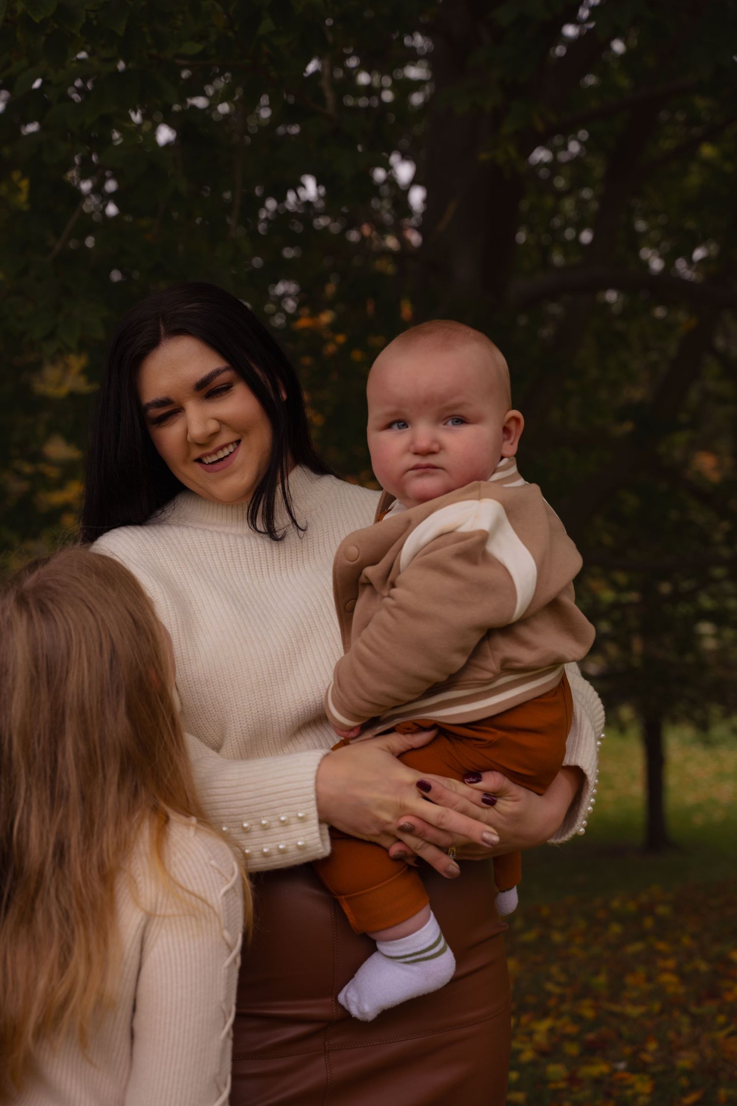Two women in cream sweaters hold a baby wearing brown clothing while enjoying an autumn day outdoors.