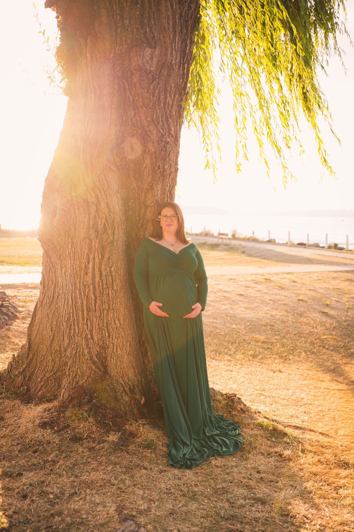 A lady in an emerald green dress poses against a sunlit willow tree creating a dreamy backlit silhouette.