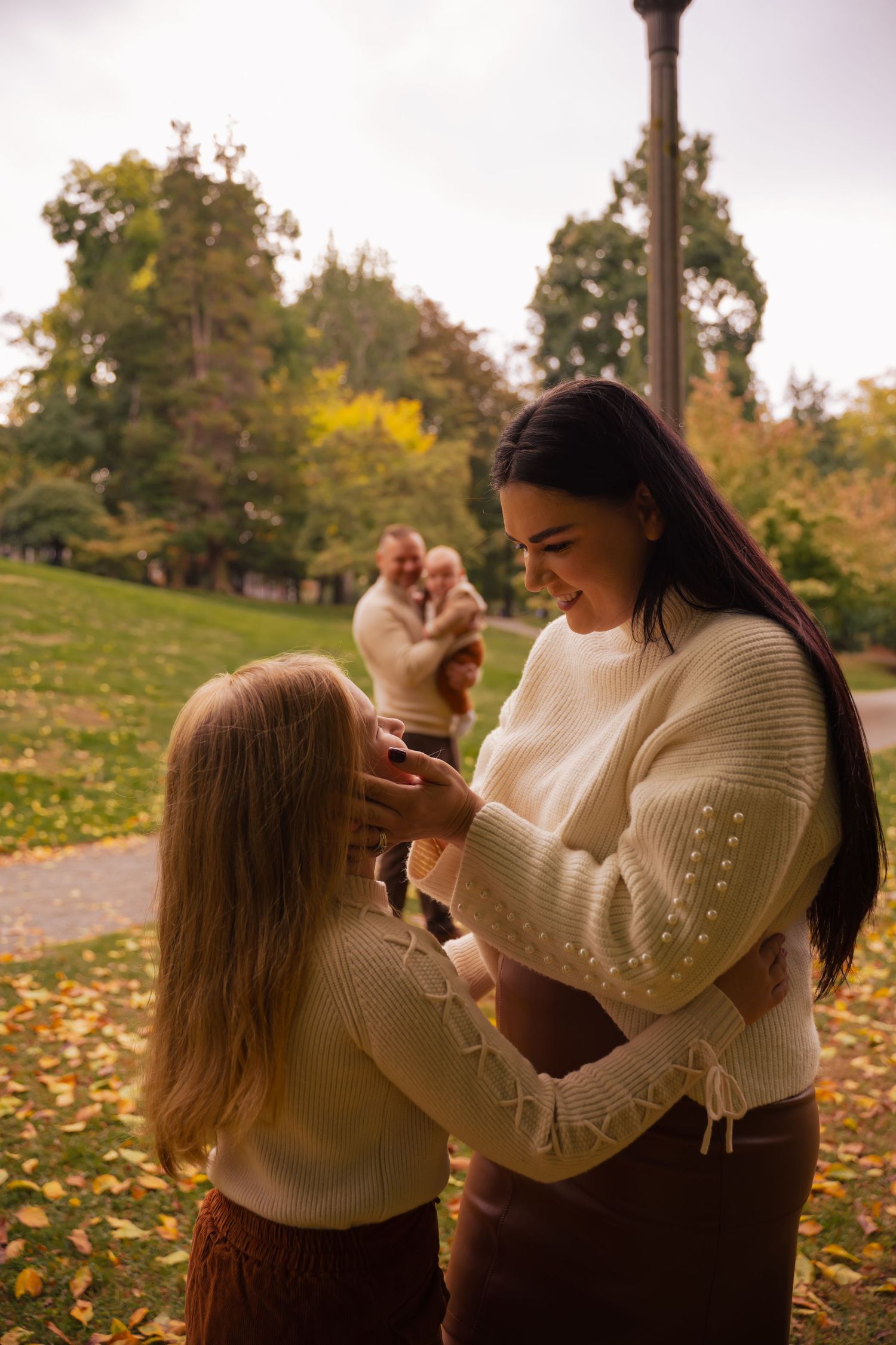 Two people in cream sweaters share a tender moment in a park during autumn with fallen leaves on the ground.