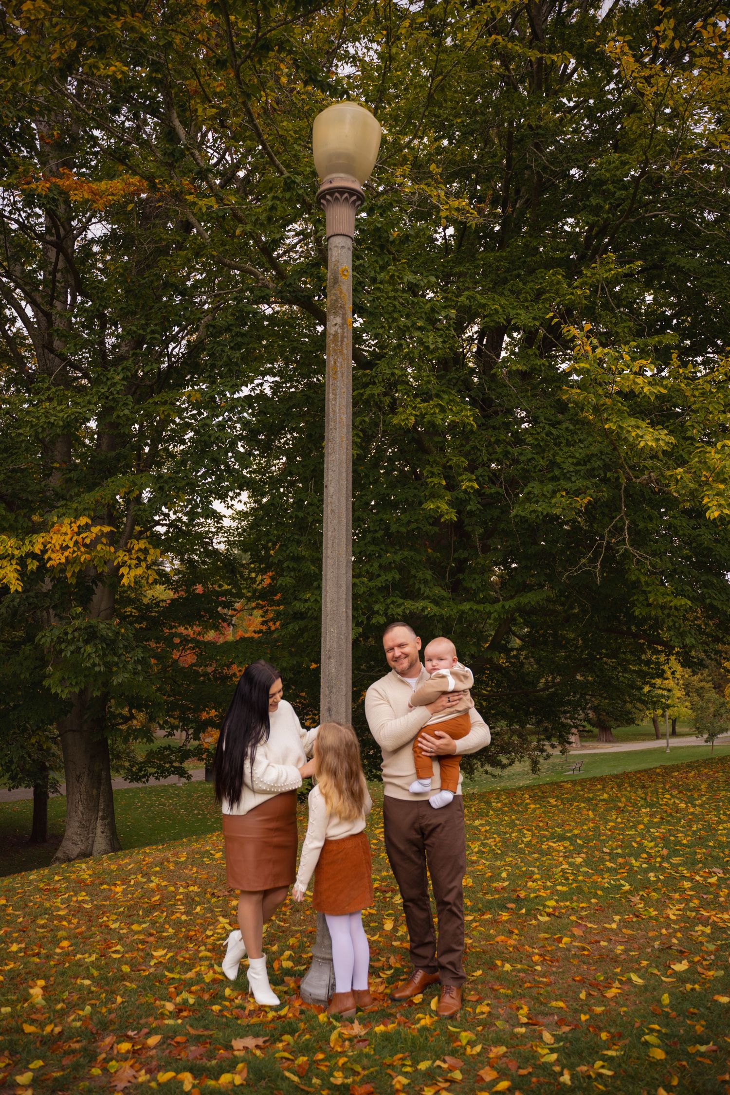Group enjoying a fall day in a park with autumn leaves scattered on grass beneath lamp posts and trees.
