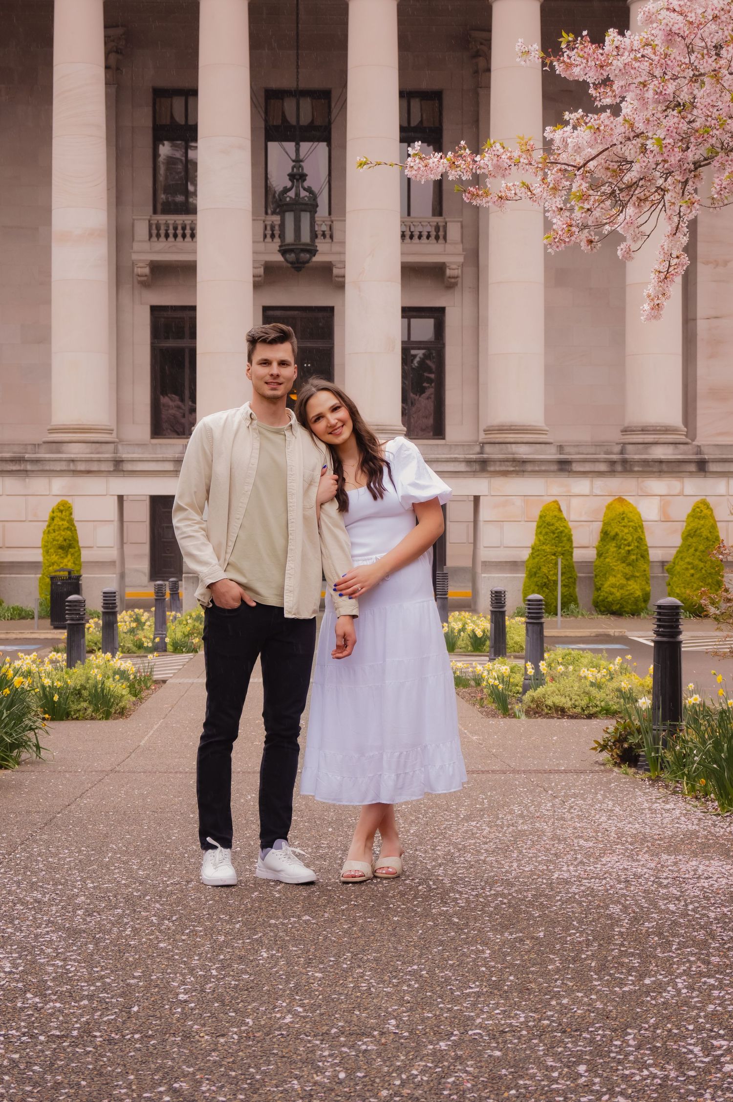 A couple poses together in front of a grand building with white columns and blooming cherry blossoms.