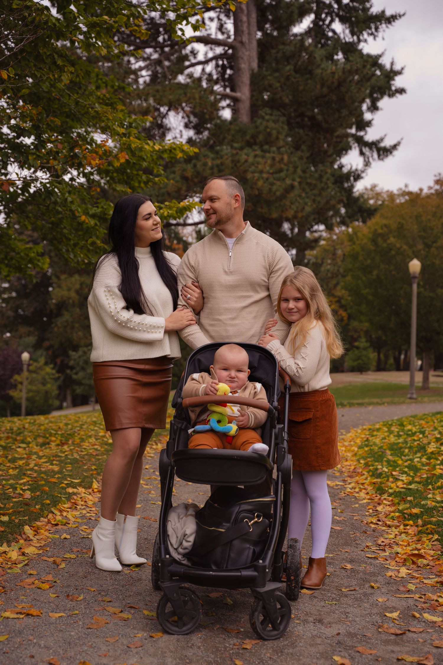 Family spending time together in autumn park with fallen leaves on ground and stroller.