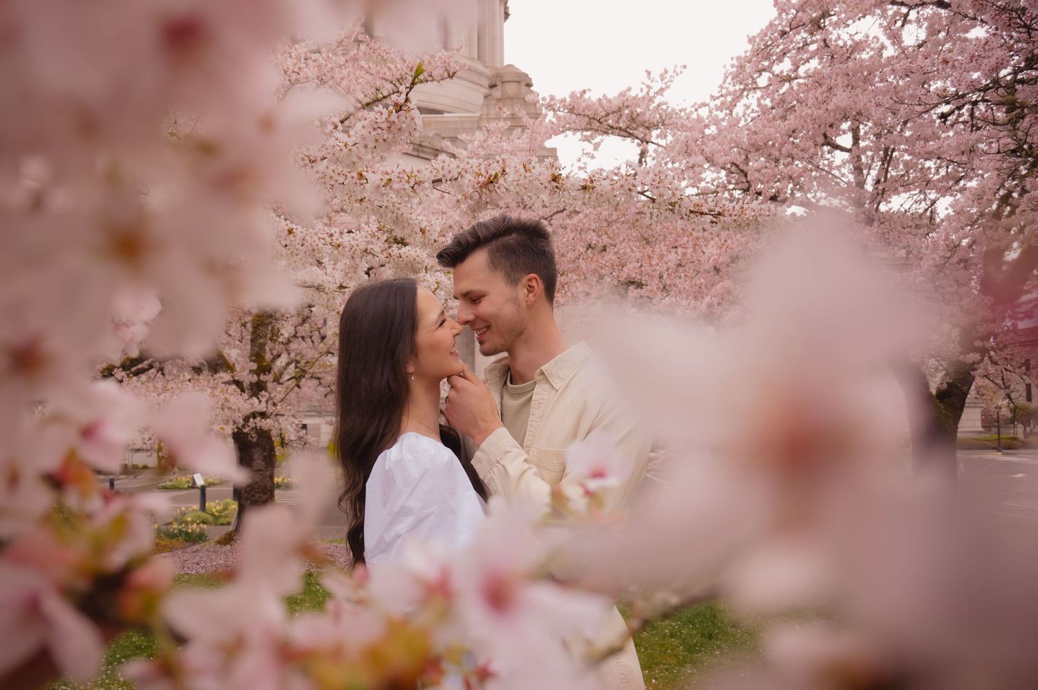A romantic couple shares an intimate moment surrounded by blooming cherry blossoms in a dreamy pink setting.