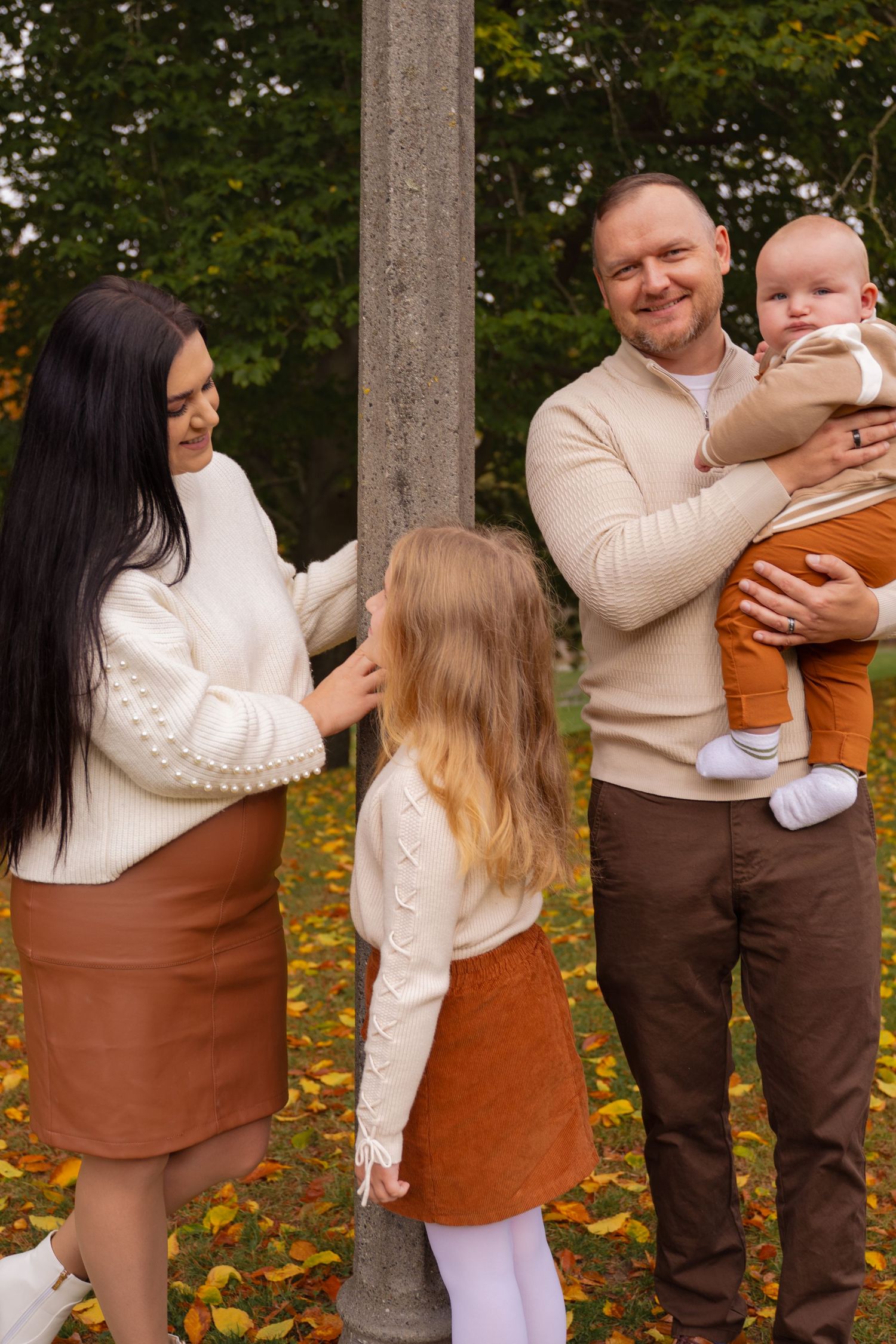 A family wearing coordinated cream and brown outfits poses together in an outdoor autumn setting.