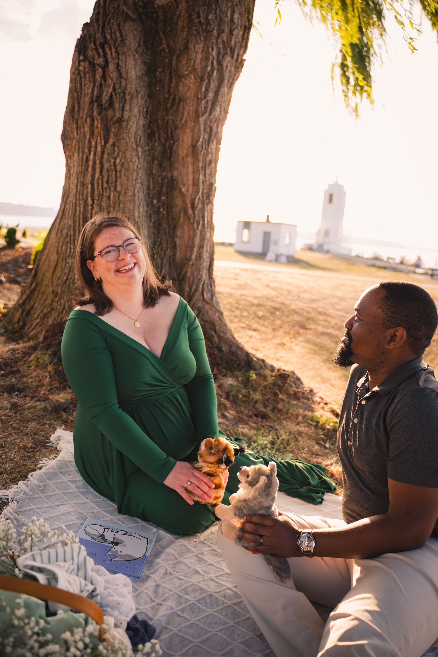 A couple shares joyful laughter while having a picnic under large trees on a sunny afternoon.