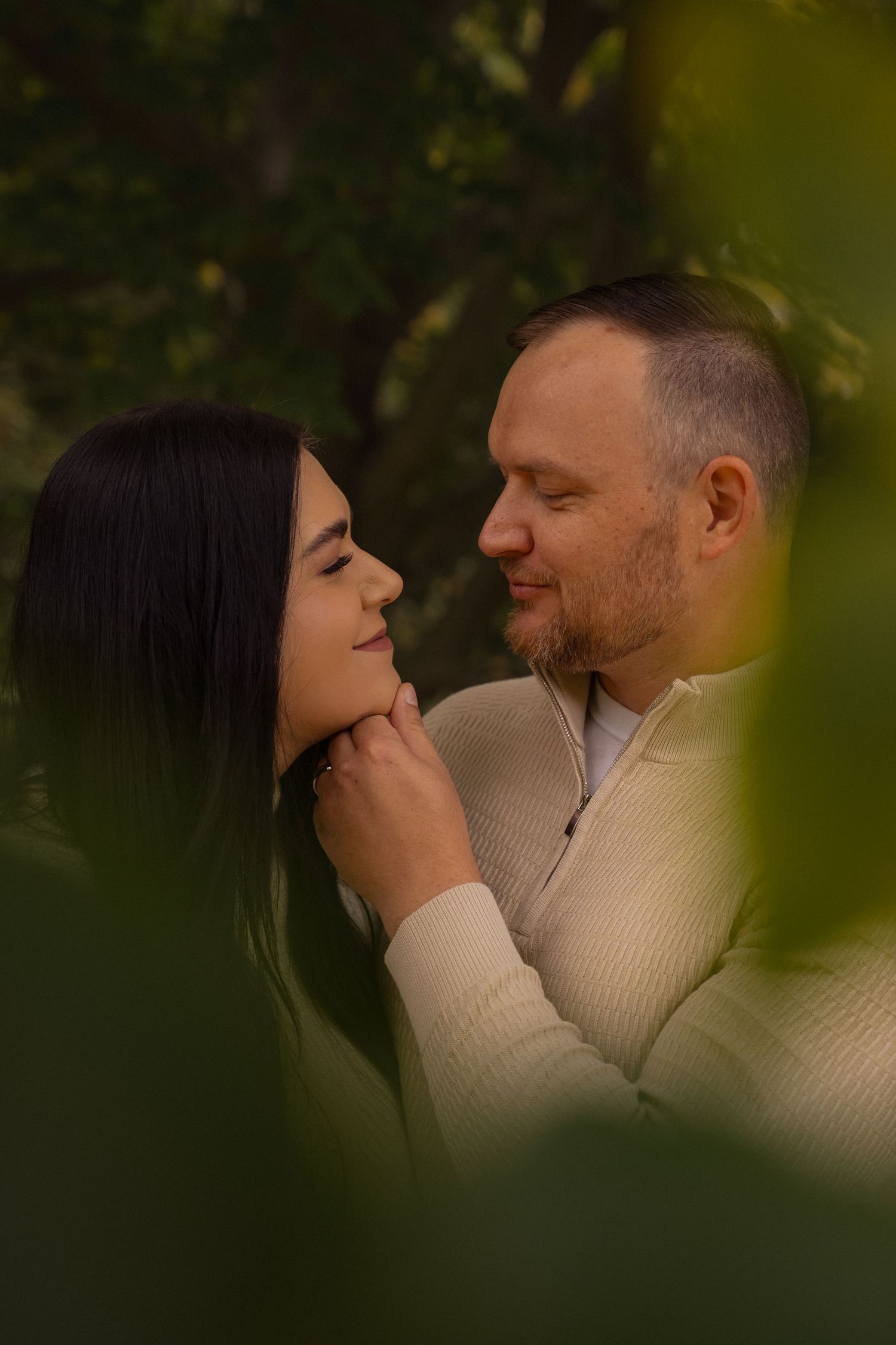 A romantic couple in cream sweaters share intimate moments through natural foliage in a series of connected photos.