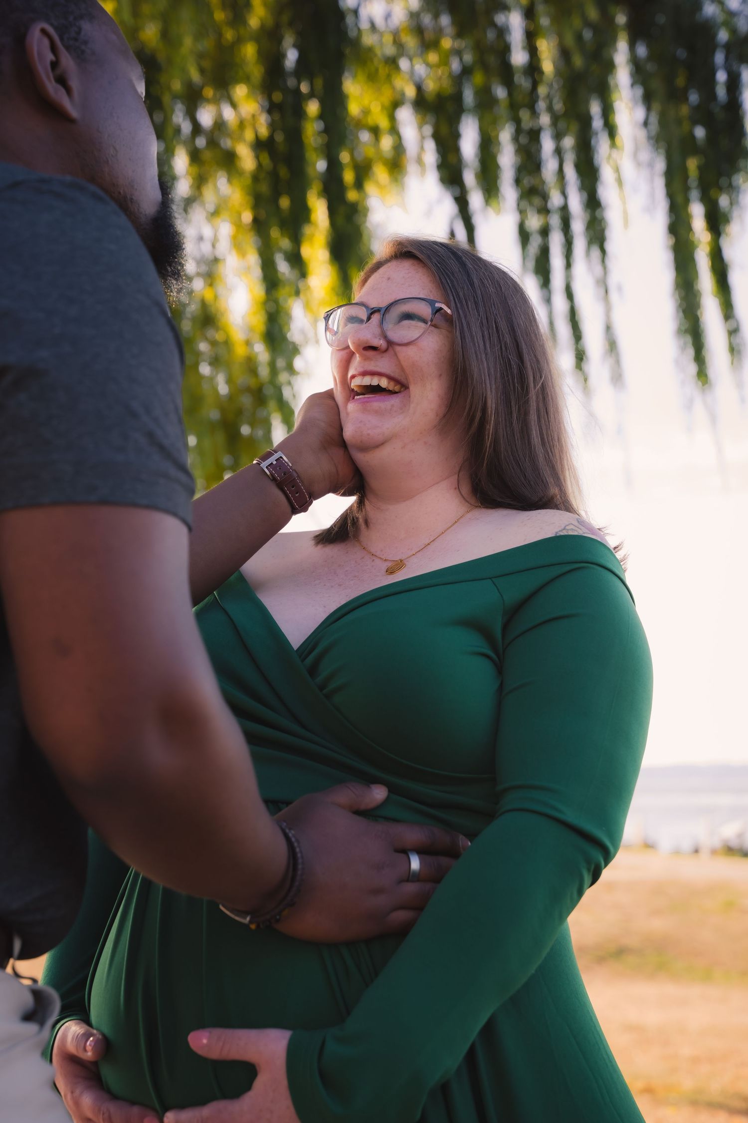 A couple shares a joyful moment outdoors with a weeping willow tree in the background during golden hour.
