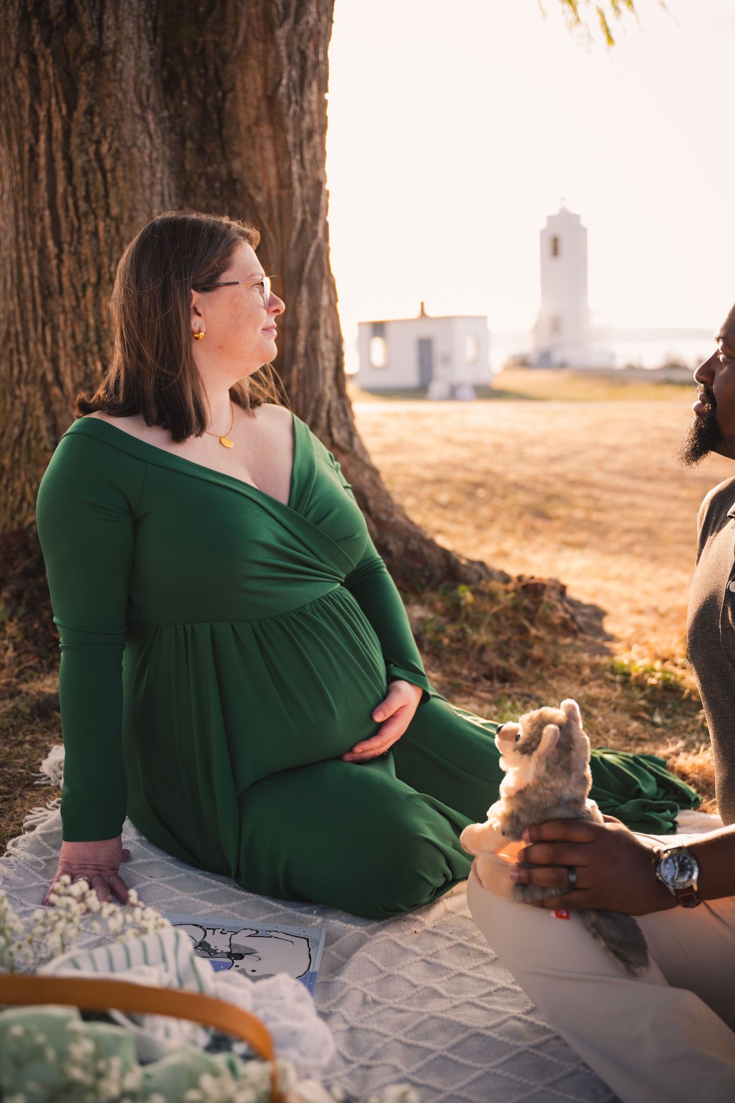 A pregnant woman in a green dress sits by a tree during a picnic with a lighthouse visible in the background.