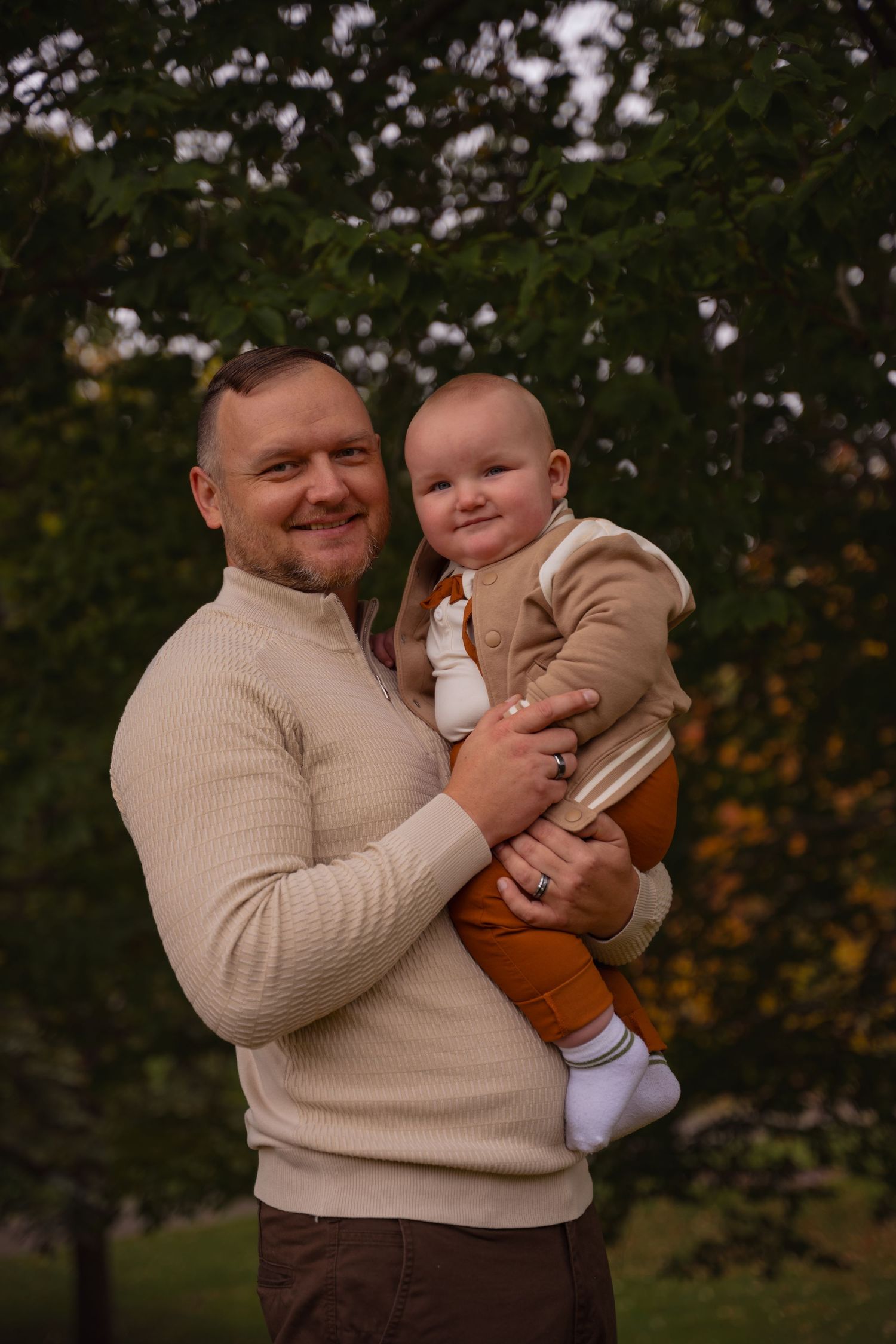 An adult and baby dressed in coordinating beige sweaters pose together outdoors against a blurred autumn background.