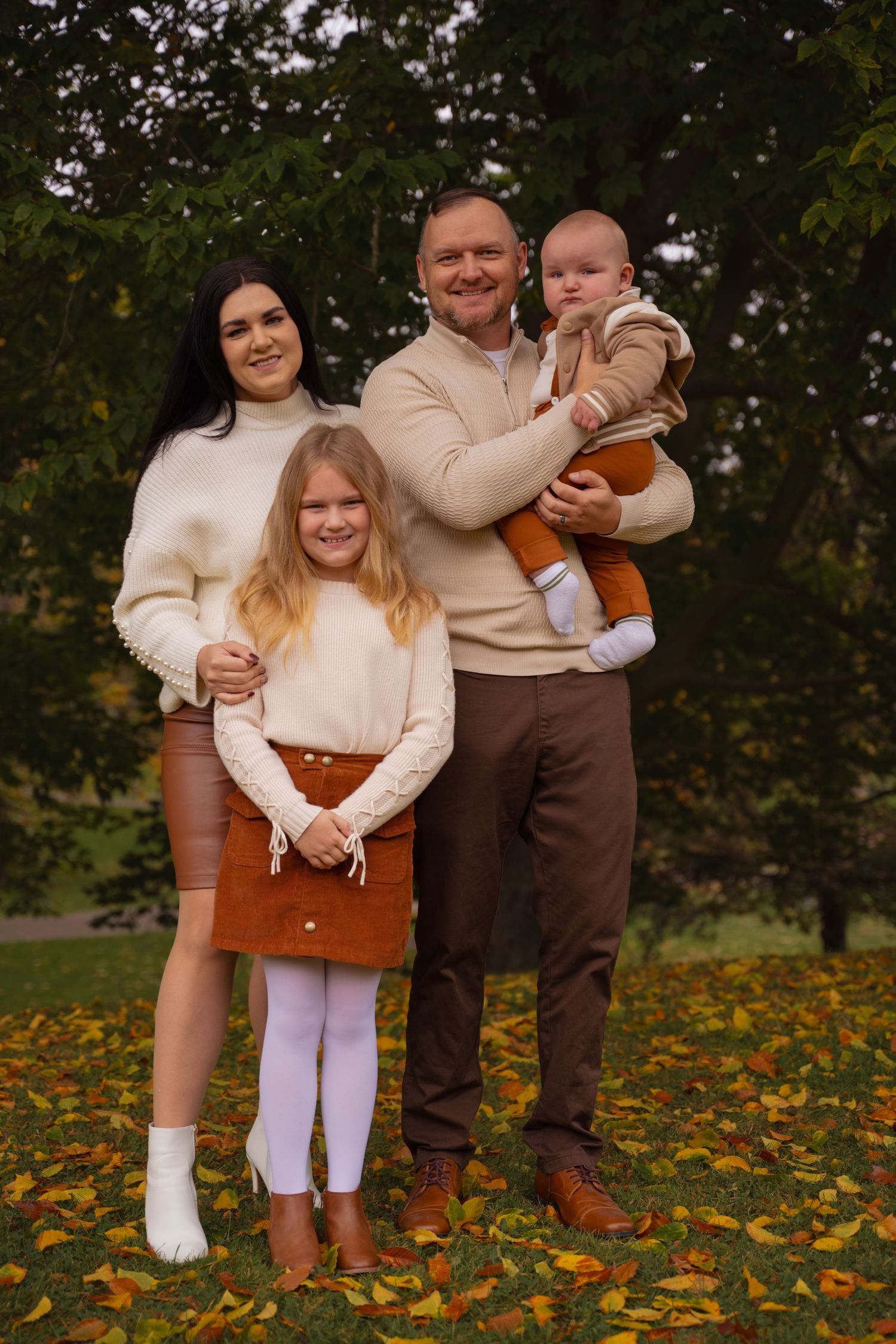 Family in coordinating cream and brown outfits poses together on fallen autumn leaves in an outdoor setting.