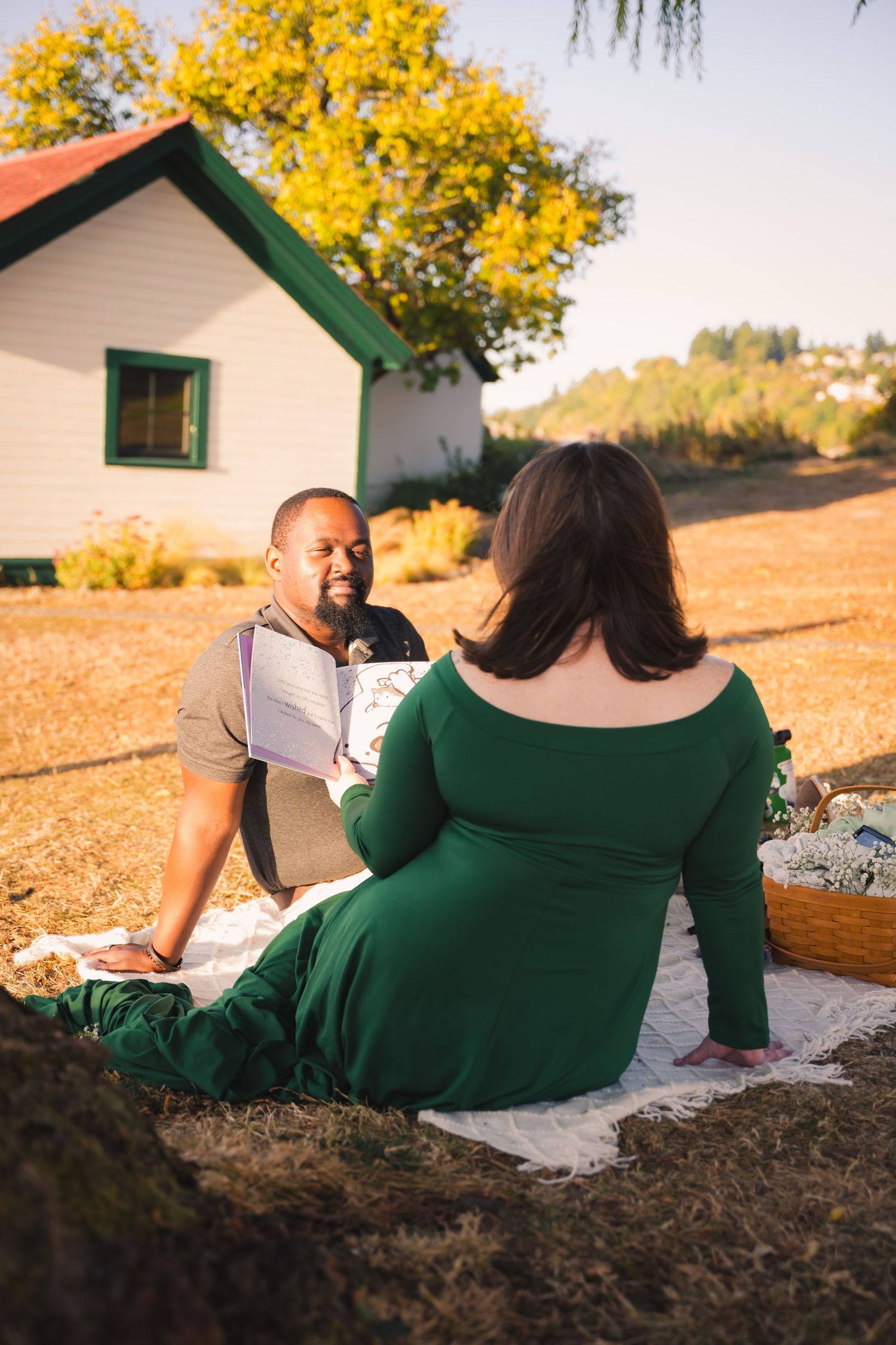 Couple relaxes together on grassy hillside during golden sunset with white building in background.