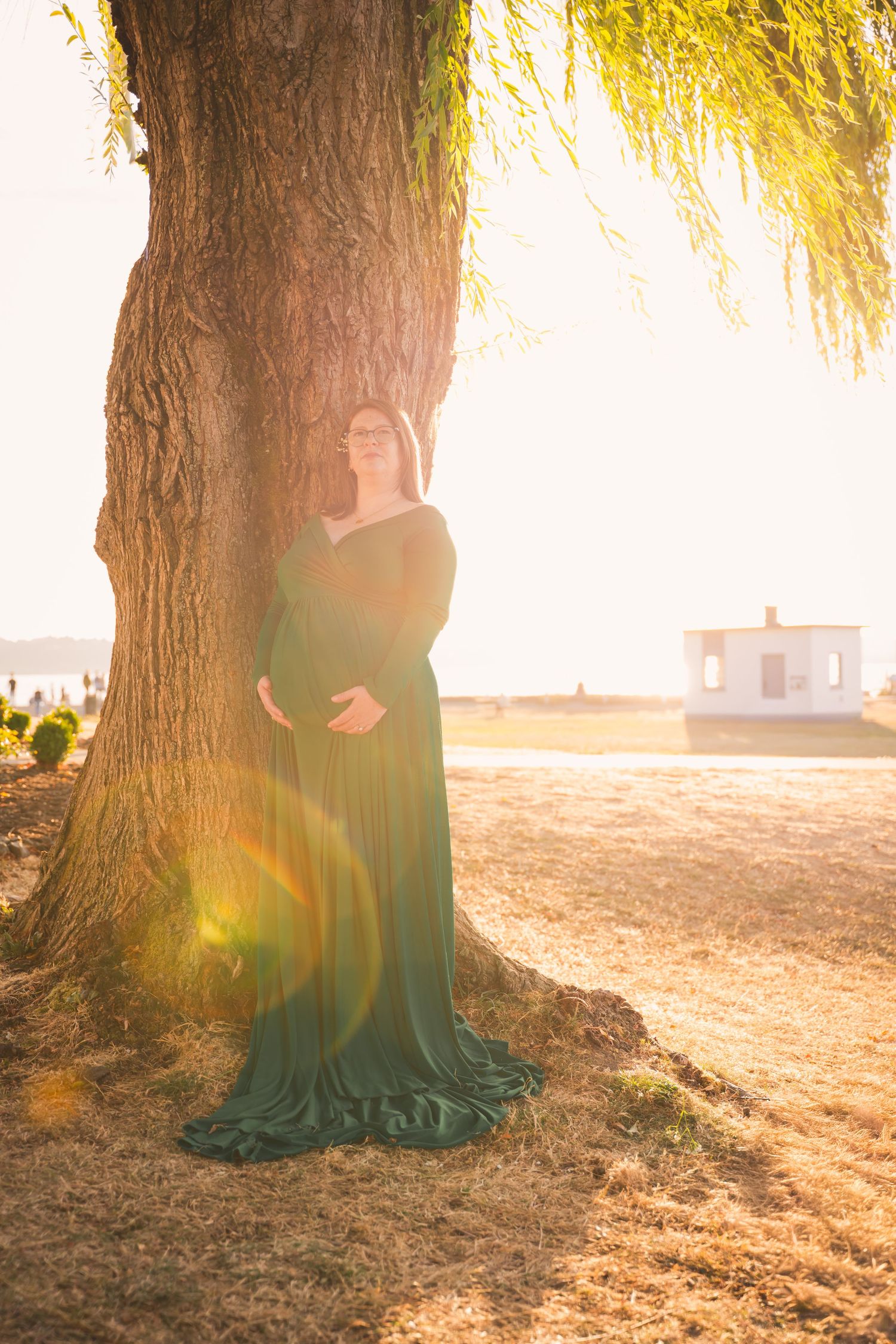 A figure in a flowing emerald green dress stands beside a large tree trunk at sunset with golden light streaming through.