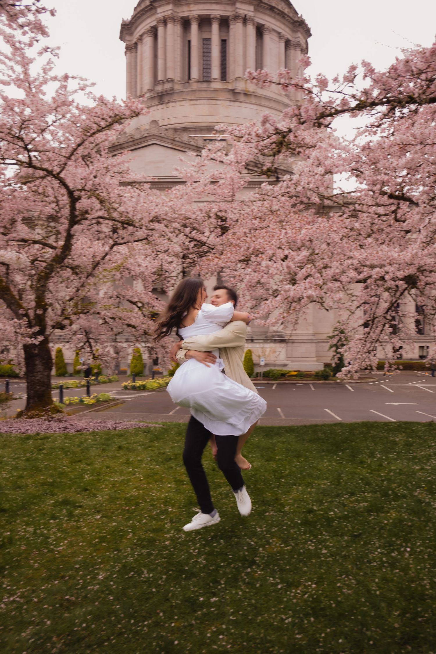 A person in a white face mask jumps and kicks against a backdrop of pink cherry blossoms and a domed building.