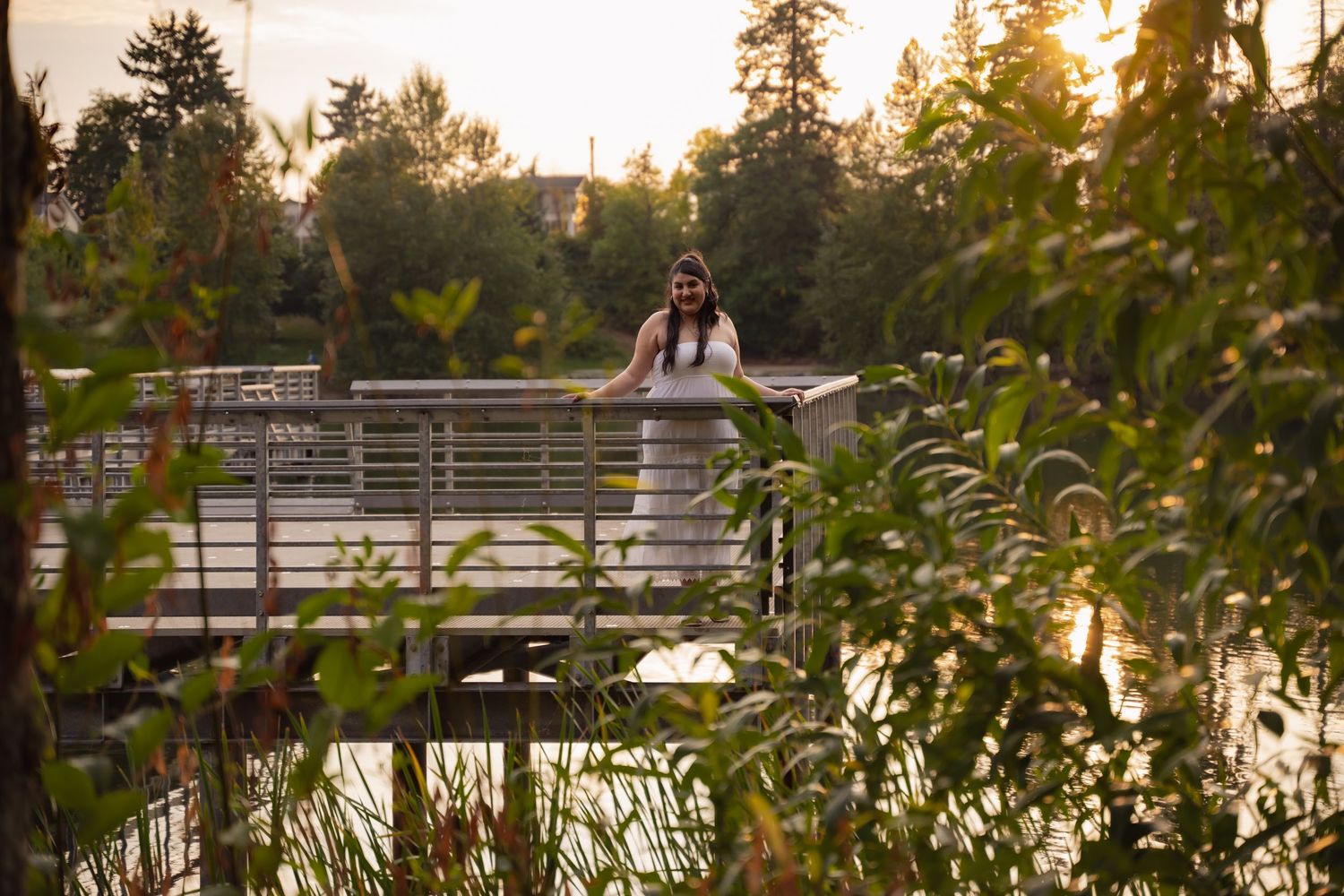 A tranquil lake surrounded by evergreen trees reflects the warm golden light of sunset through a wooden bridge.