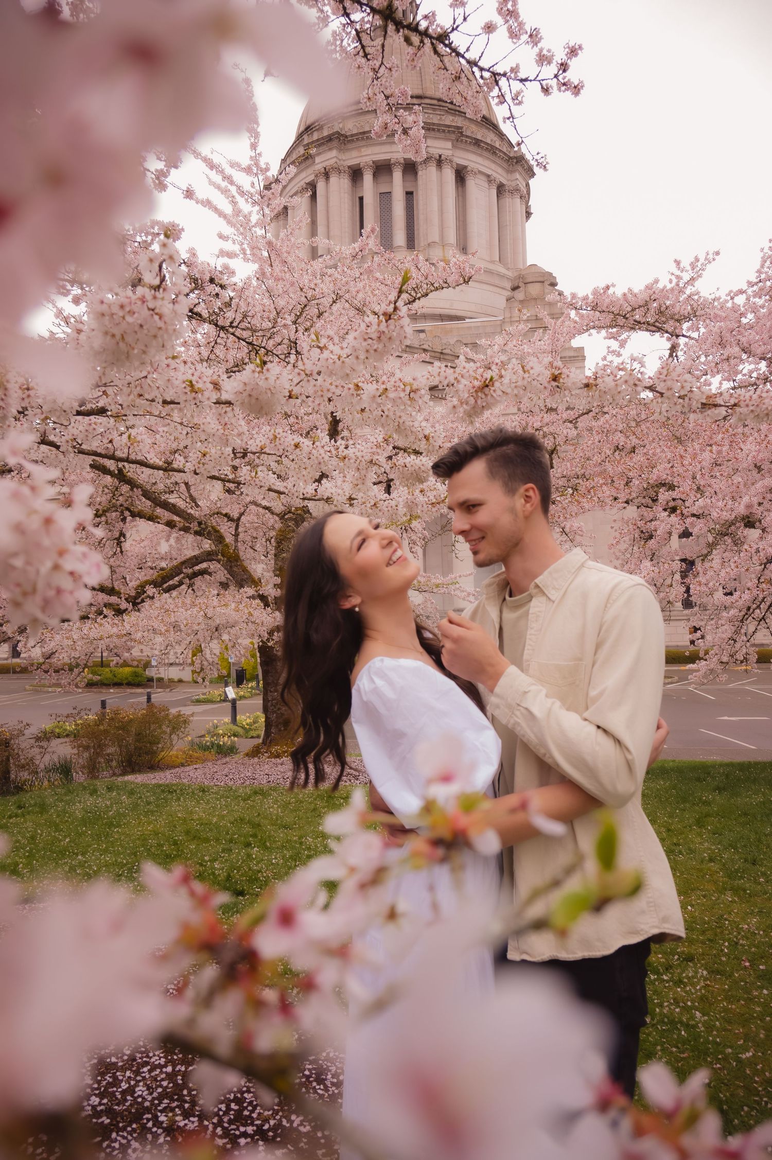 A romantic couple embraces under blooming cherry blossoms with a domed capitol building in the background.