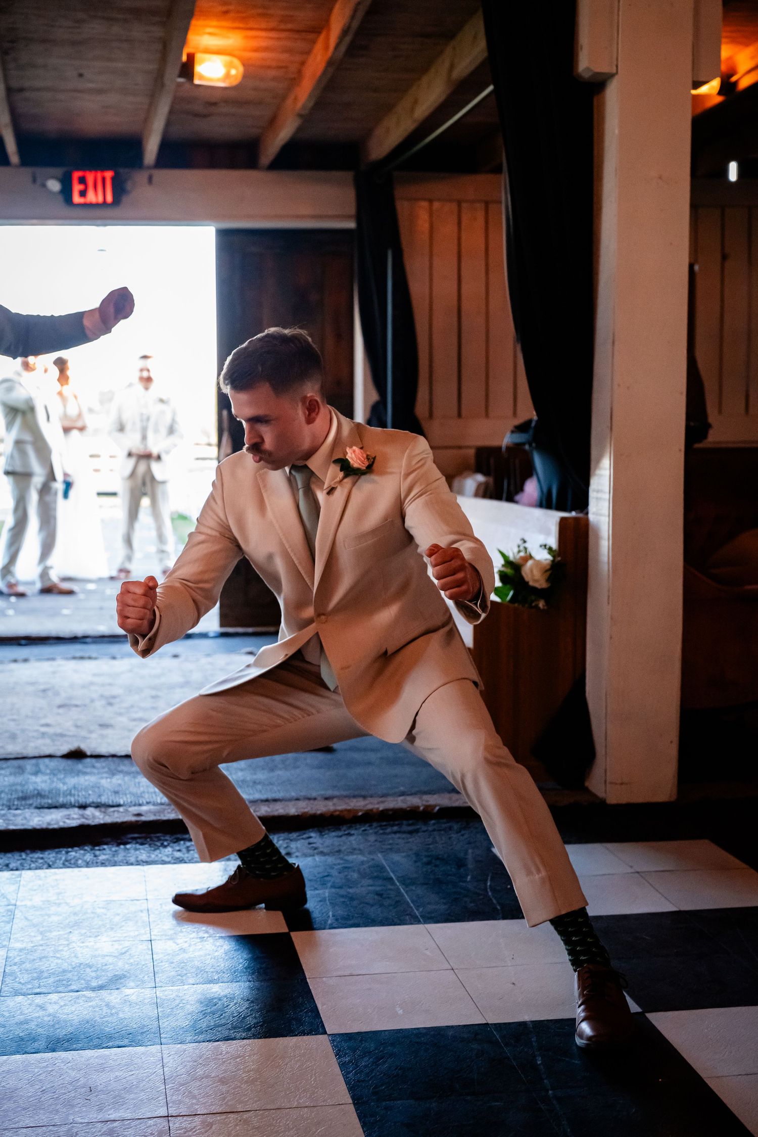 Groom in beige suit performing choreographed dance moves indoors.