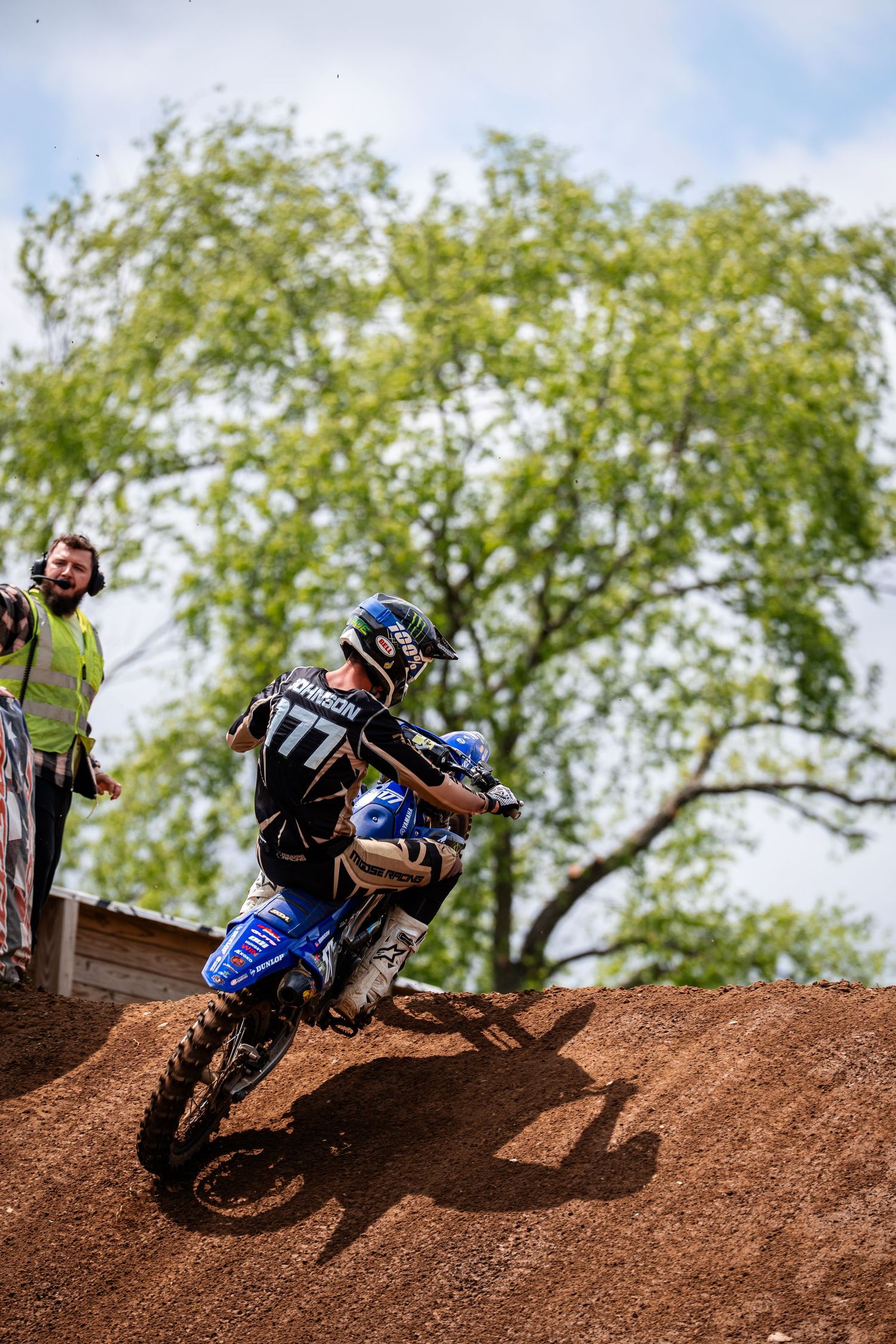 Motocross rider navigates dirt jump on blue motorcycle against green tree background.