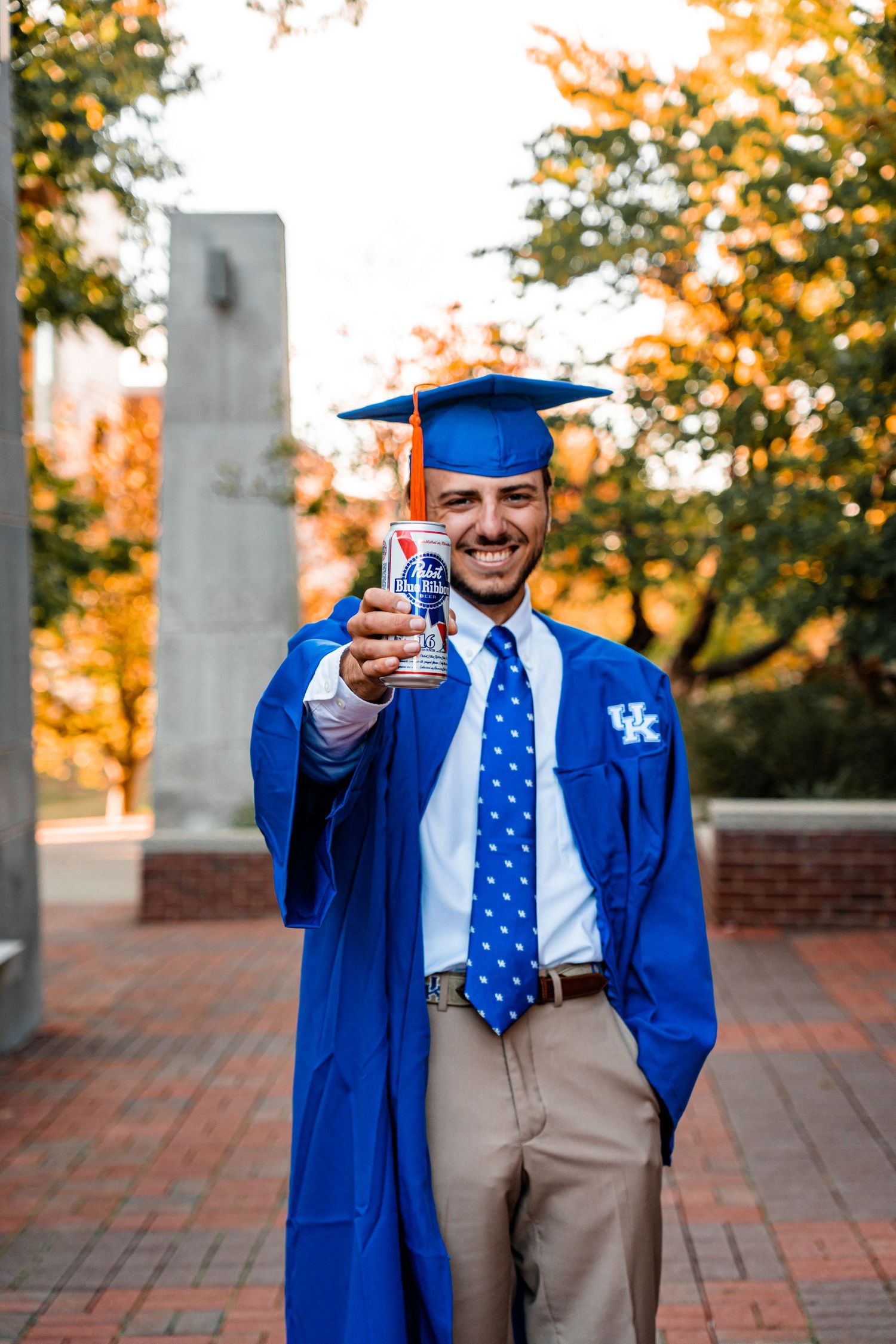 Graduate in blue UK cap and gown celebrates with diploma on campus.