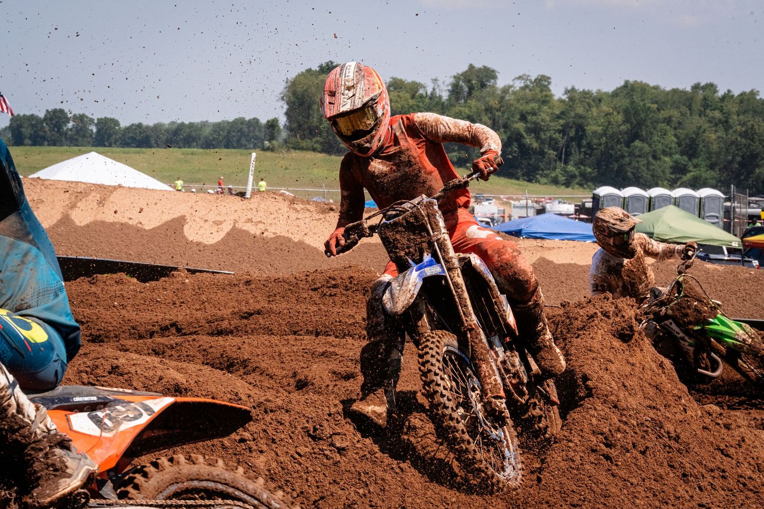 A motocross rider in protective gear races through a dirt track surrounded by nature.