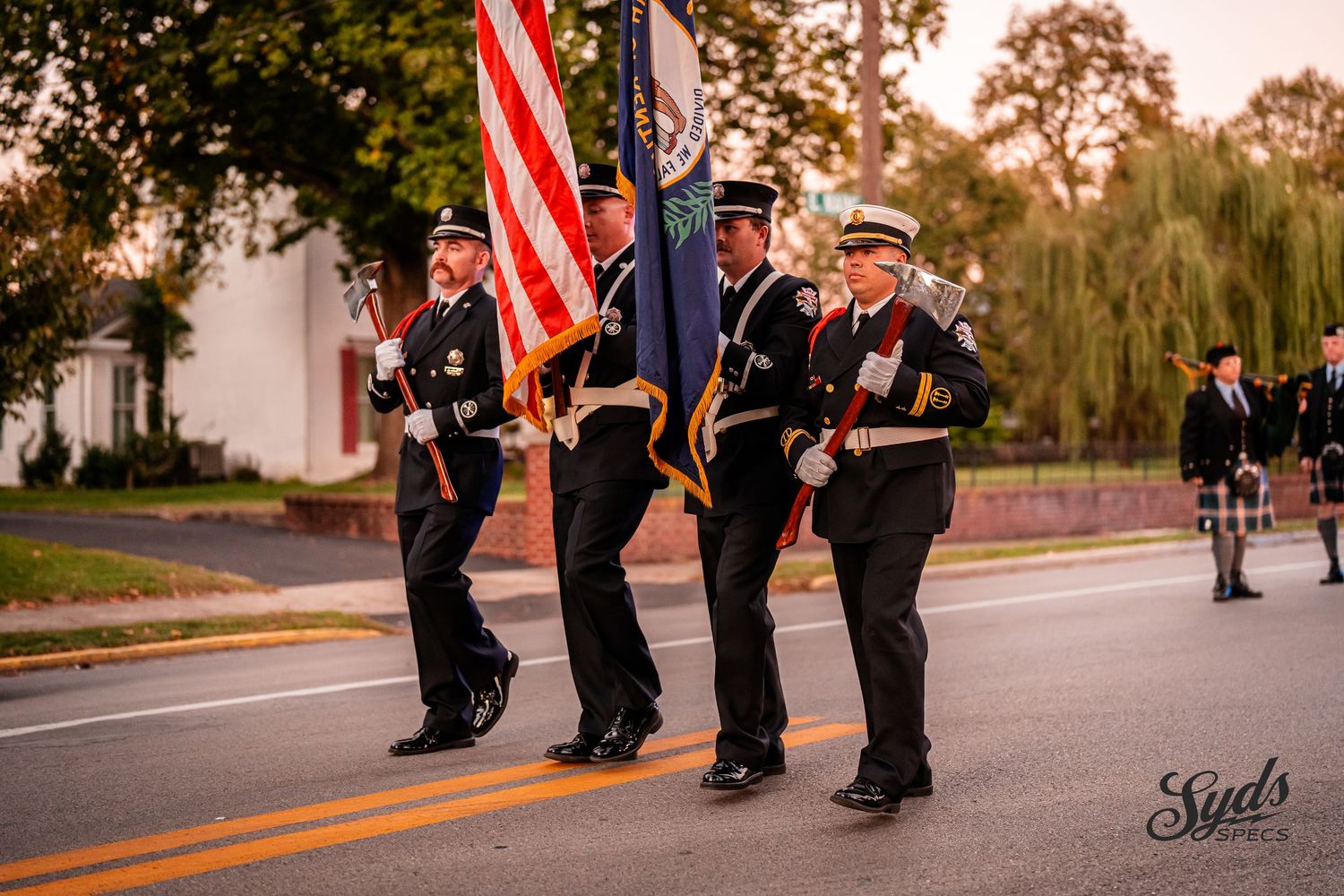 Firefighters march in formation along a street at sunset with American flags flying overhead.