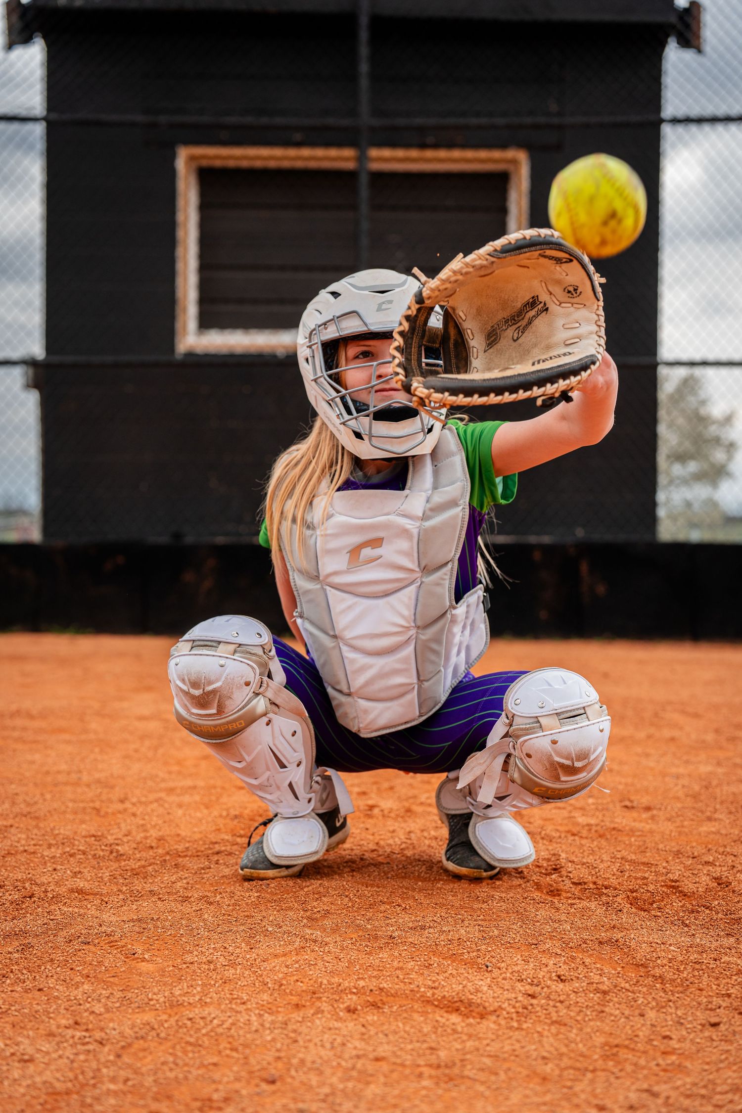 Softball catcher in protective gear crouches to catch ball on dirt field.