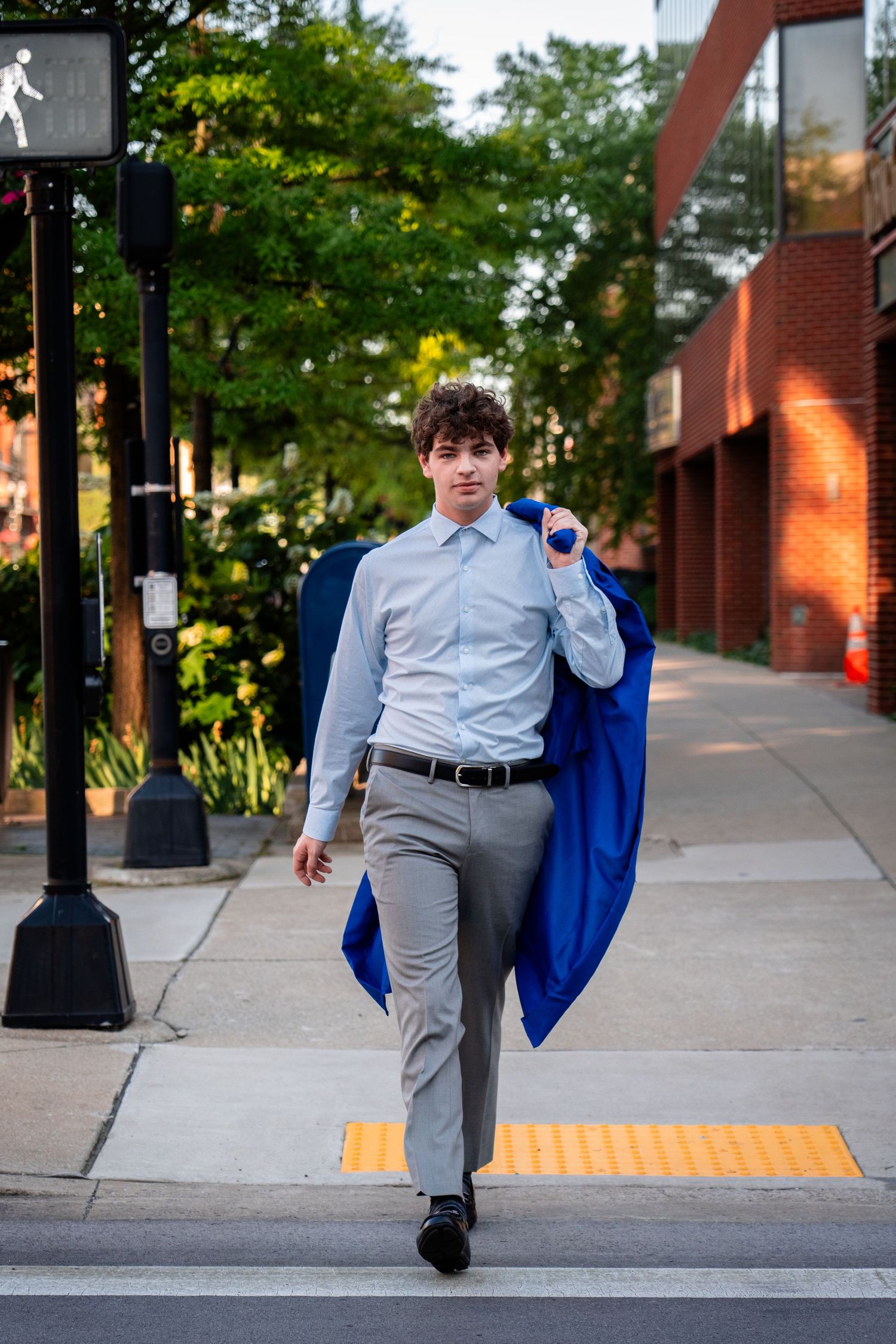 Graduate in gray suit carrying blue graduation gown while walking outdoors.