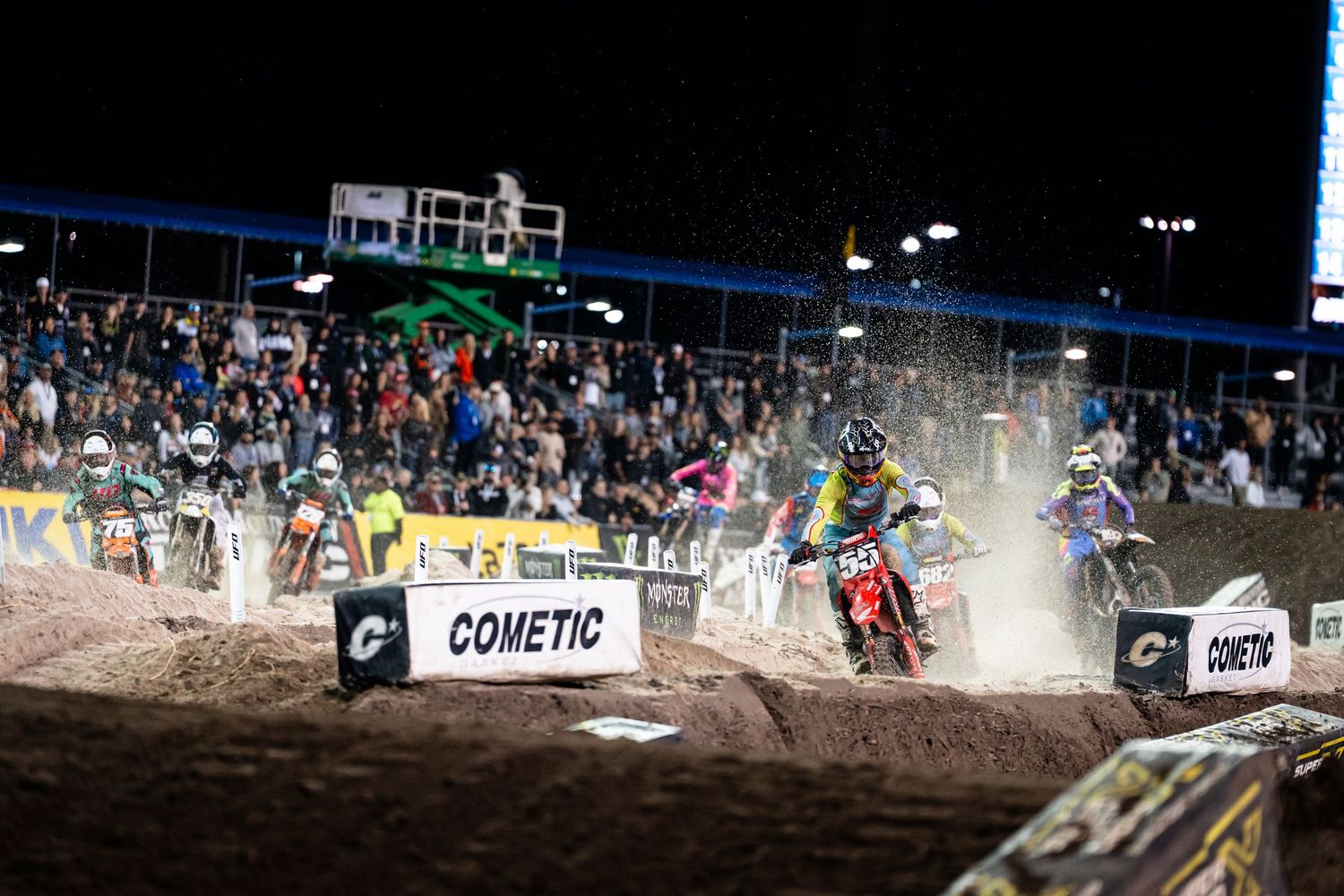 Motocross riders racing through dirt track under stadium lights.