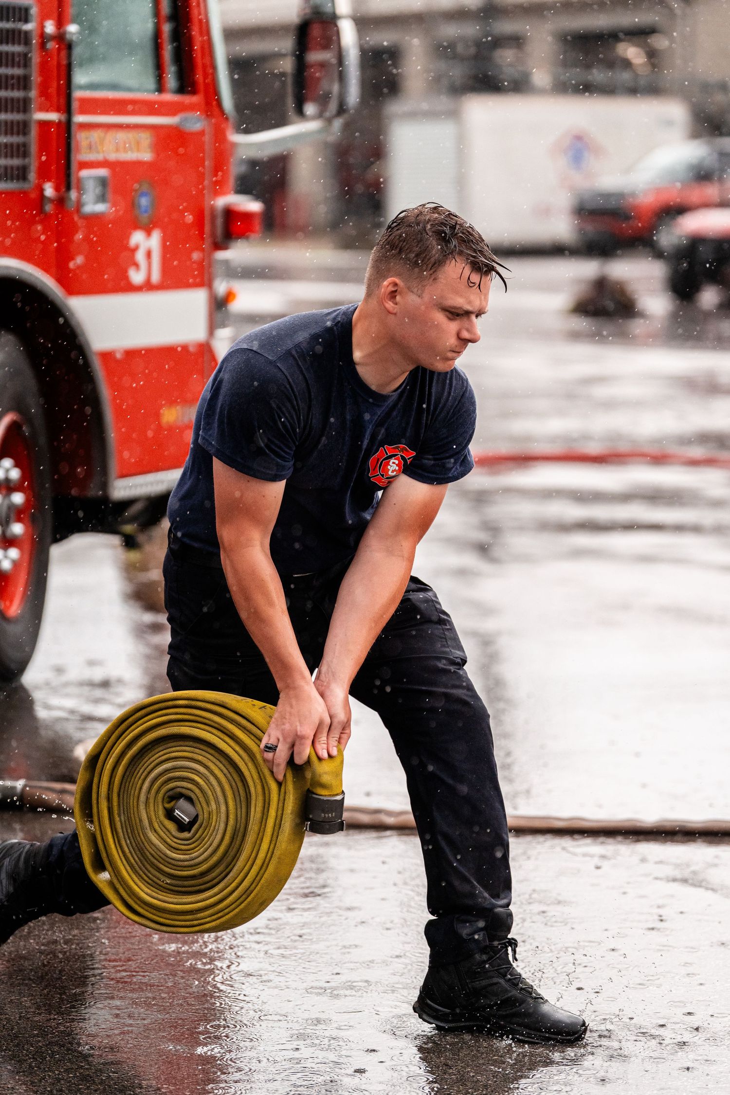 Firefighter rolls up a yellow fire hose on wet pavement during equipment maintenance training.