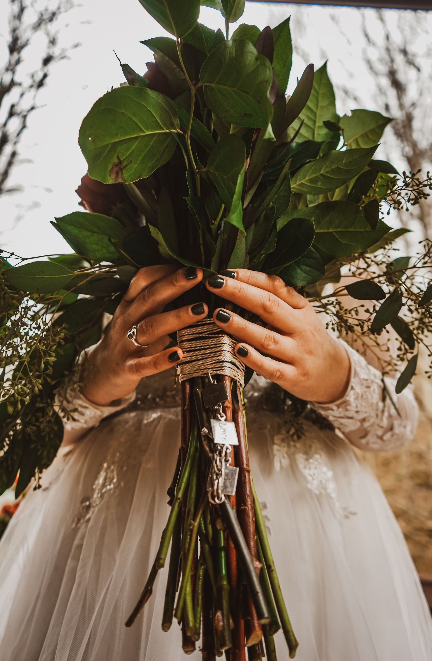 Close-up of hands holding wedding bouquet with greenery against white dress.