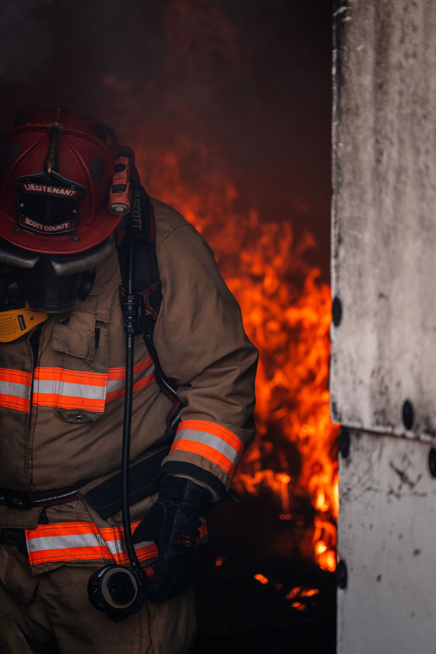 Firefighter in protective gear stands against backdrop of intense flames during nighttime operation.