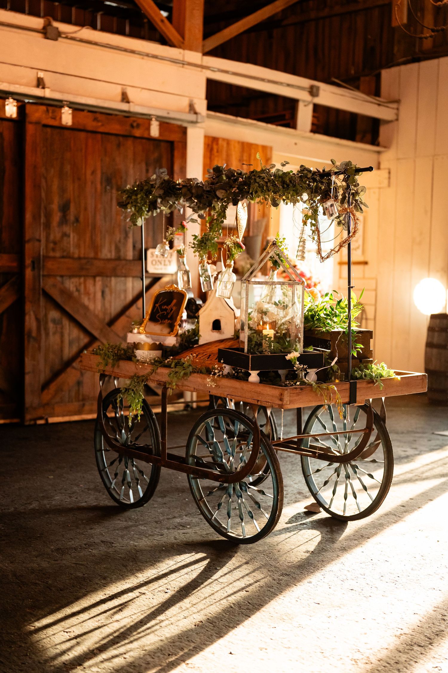 Vintage wooden cart decorated with greenery and lanterns creates a rustic wedding display in a barn setting.