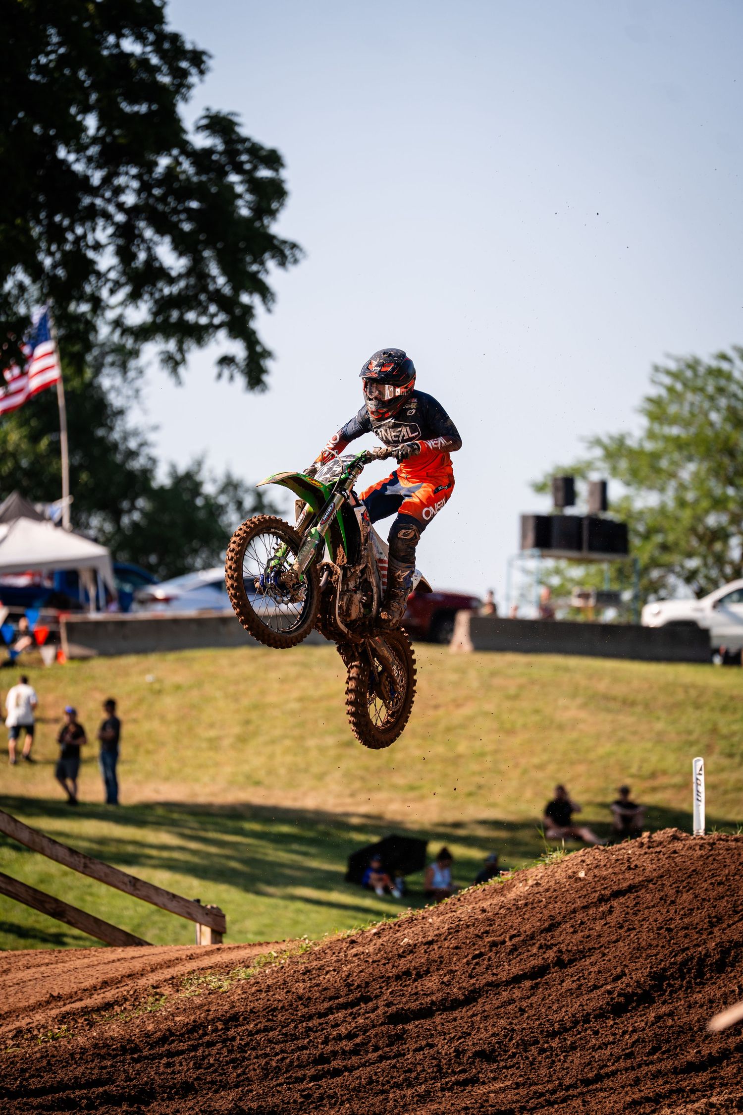 Dirt bike rider soars over jump on motocross track with American flag visible in background.