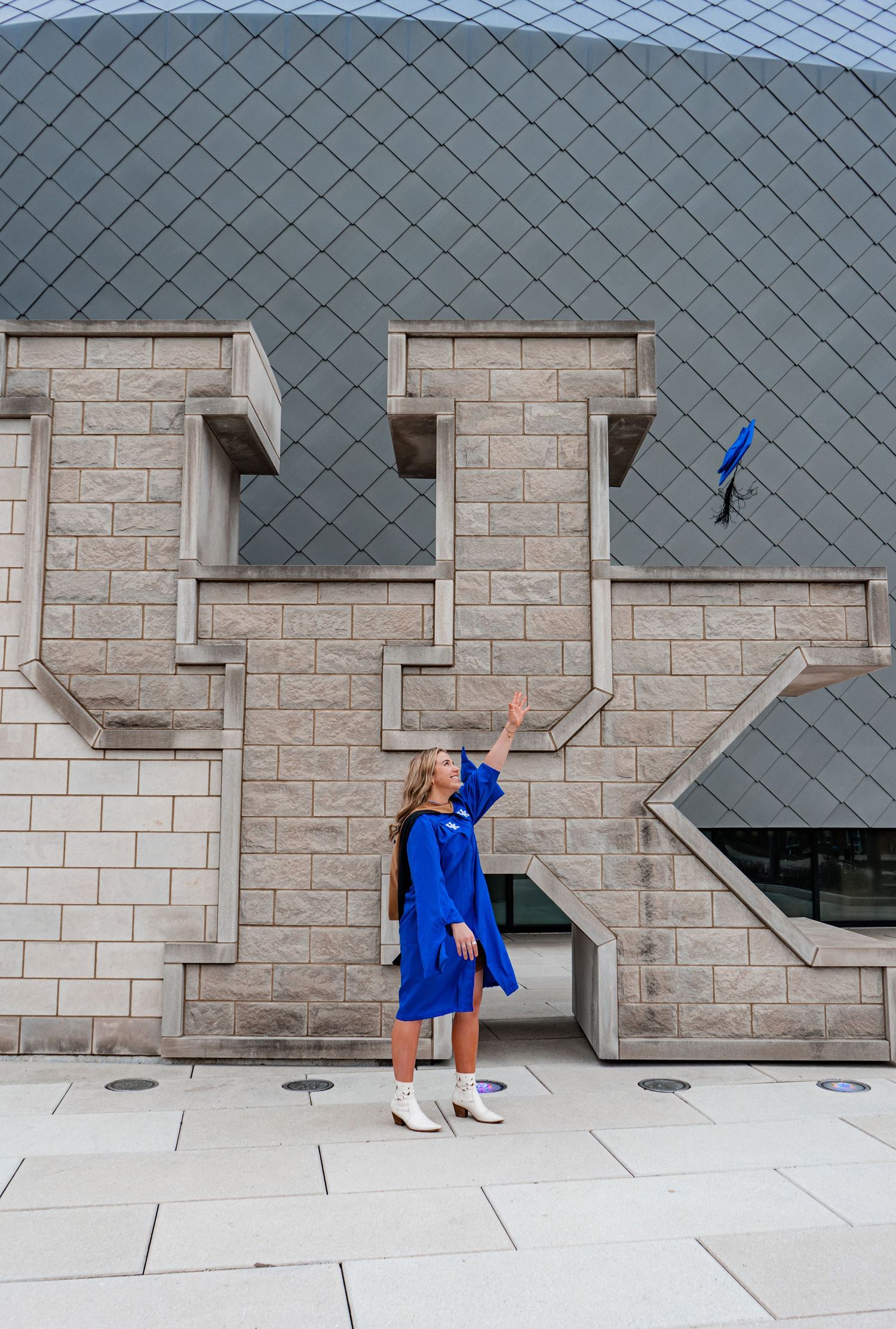 Graduate in blue cap and gown throws graduation cap against modern architectural backdrop.