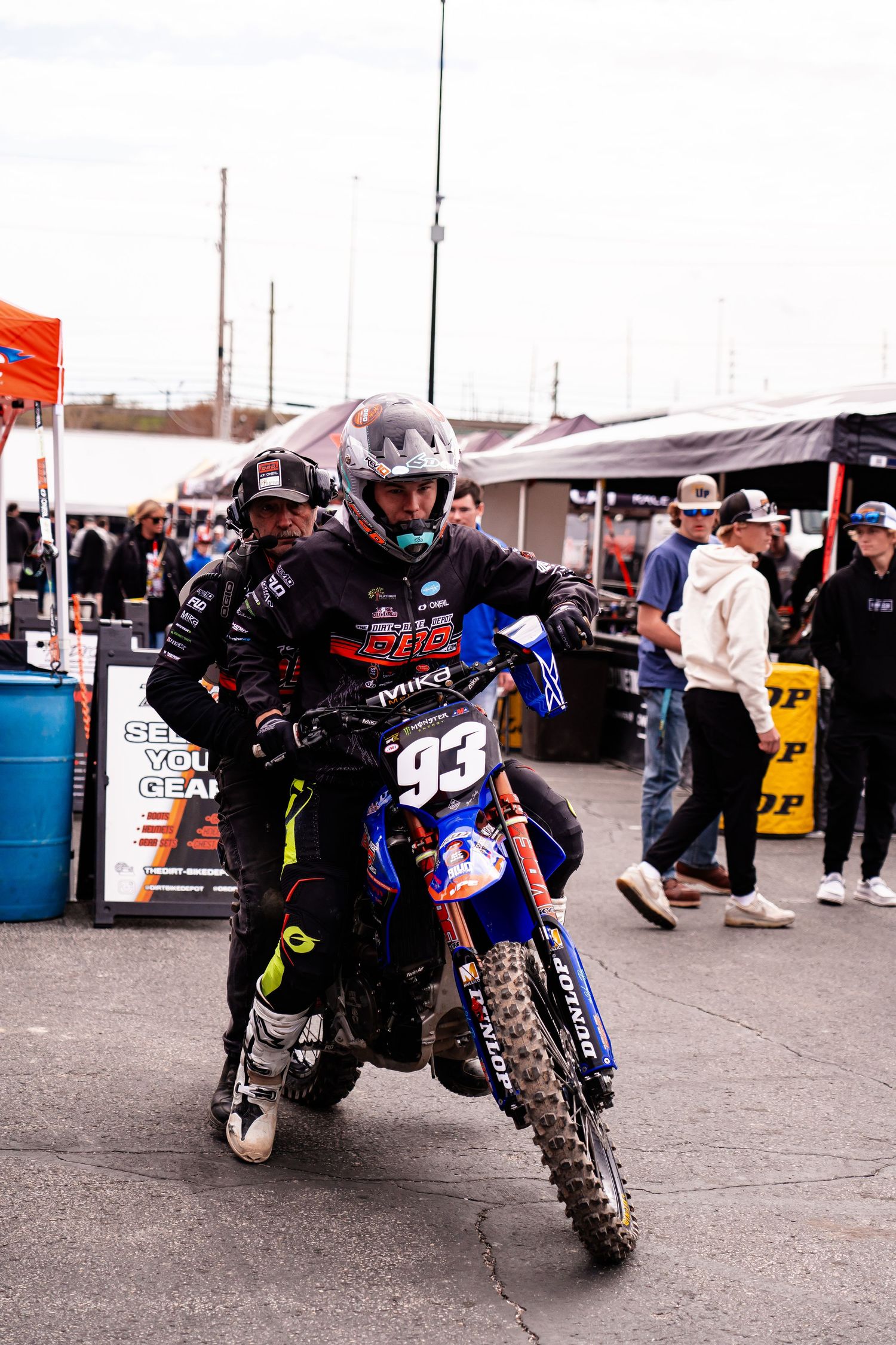 Dirt bike rider performing stunts and wheelies in a parking lot with spectators watching.