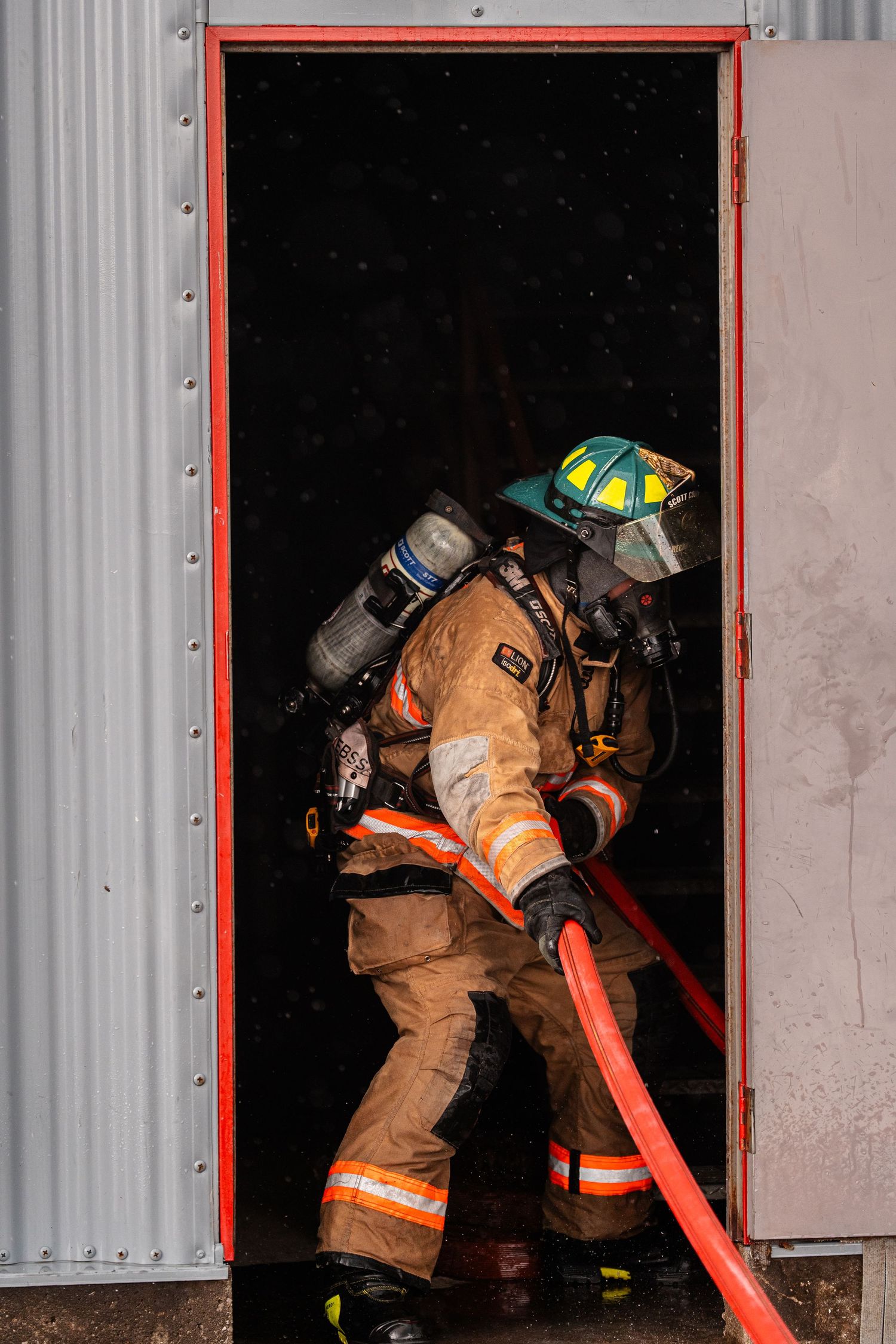 A firefighter in full protective gear pulls a hose through a doorway during an operation.