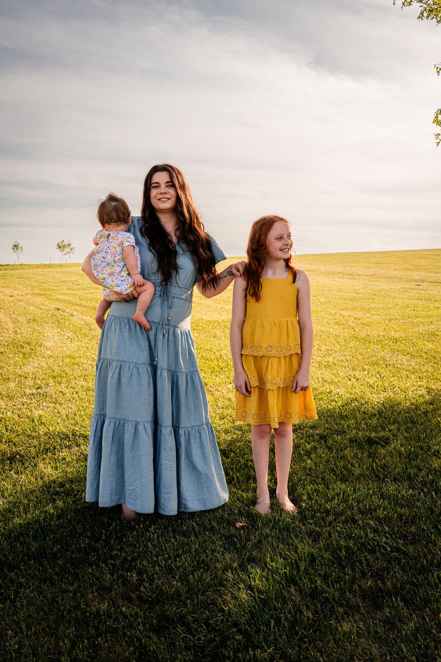 Three people in coordinating blue and yellow dresses pose together in a sunny meadow.