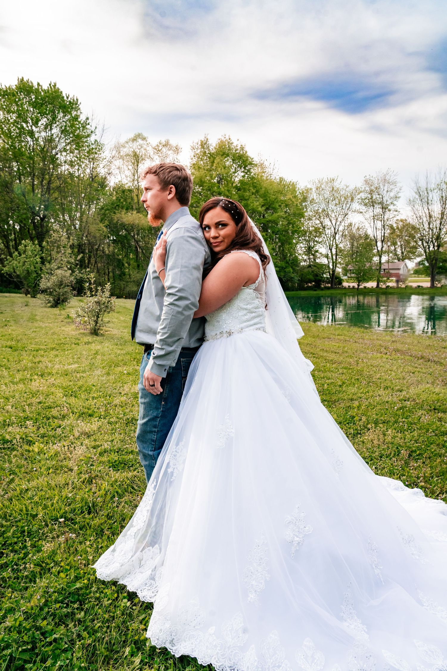 Bride in flowing white gown and groom pose back-to-back by lakeside.