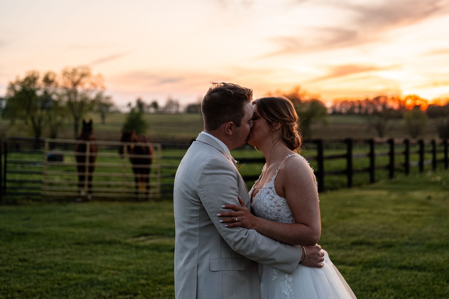 Newlyweds share an intimate moment at sunset in a pastoral field with wooden fences.