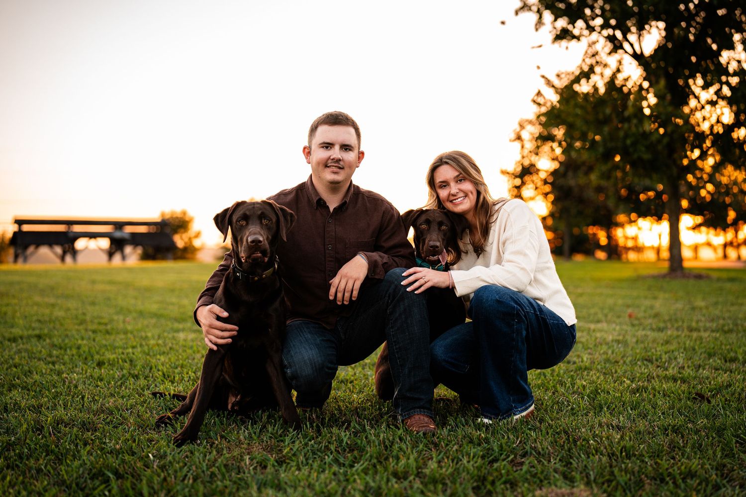 A couple poses with their two black dogs in a sunset-lit field.