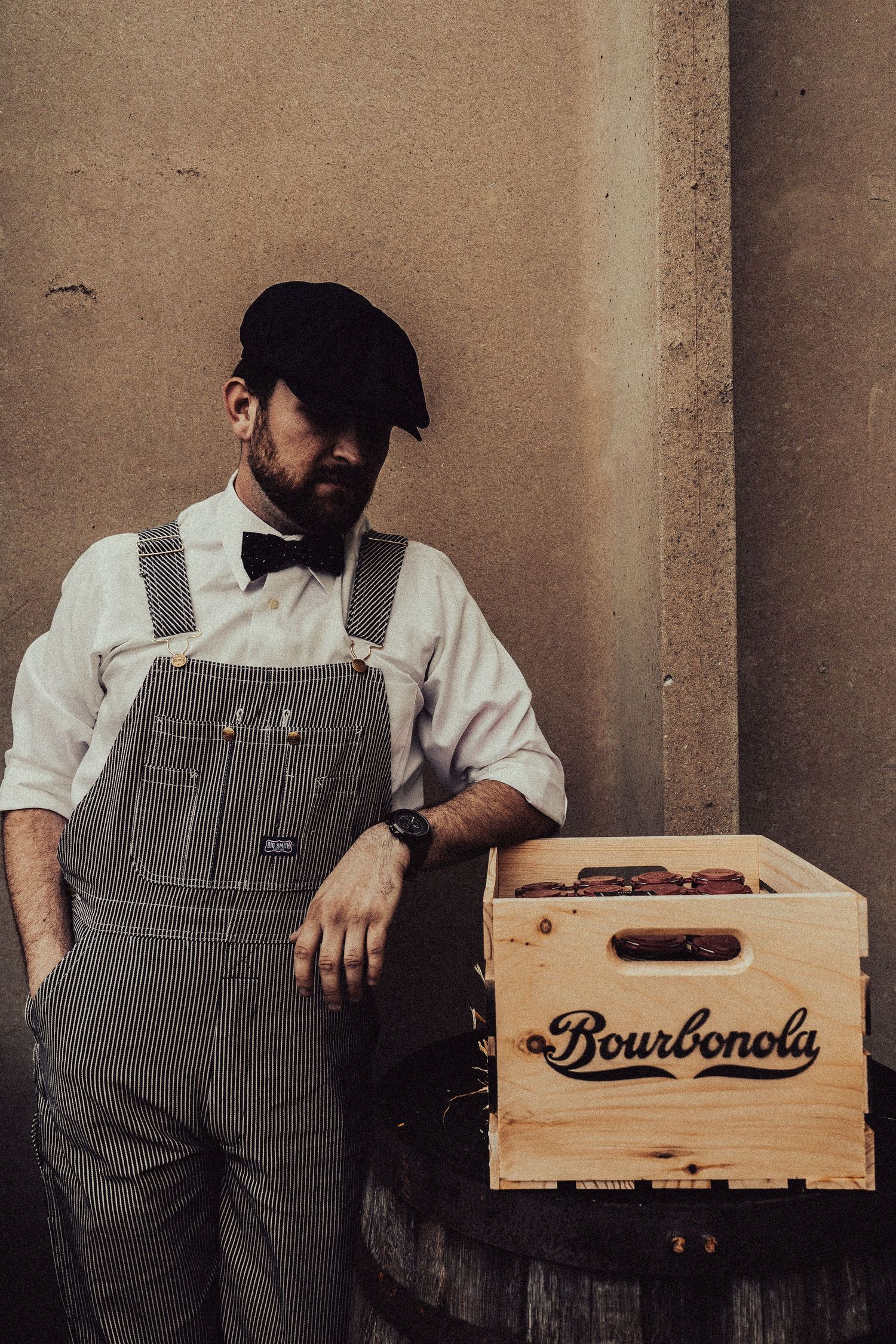 Worker in overalls sitting with wooden Bourbonola crate in industrial setting.