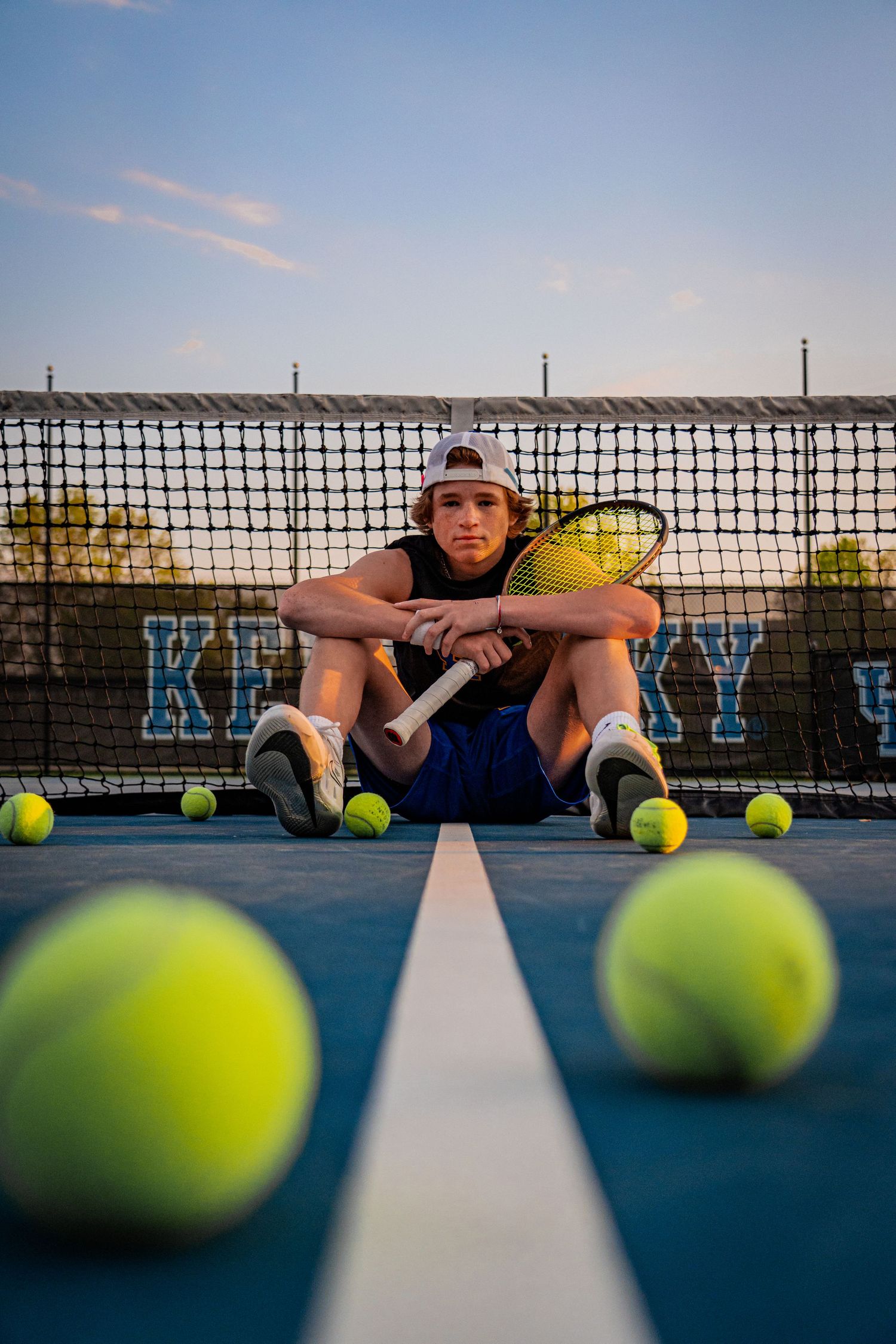 Tennis player rests against net with tennis balls scattered on blue court at sunset.