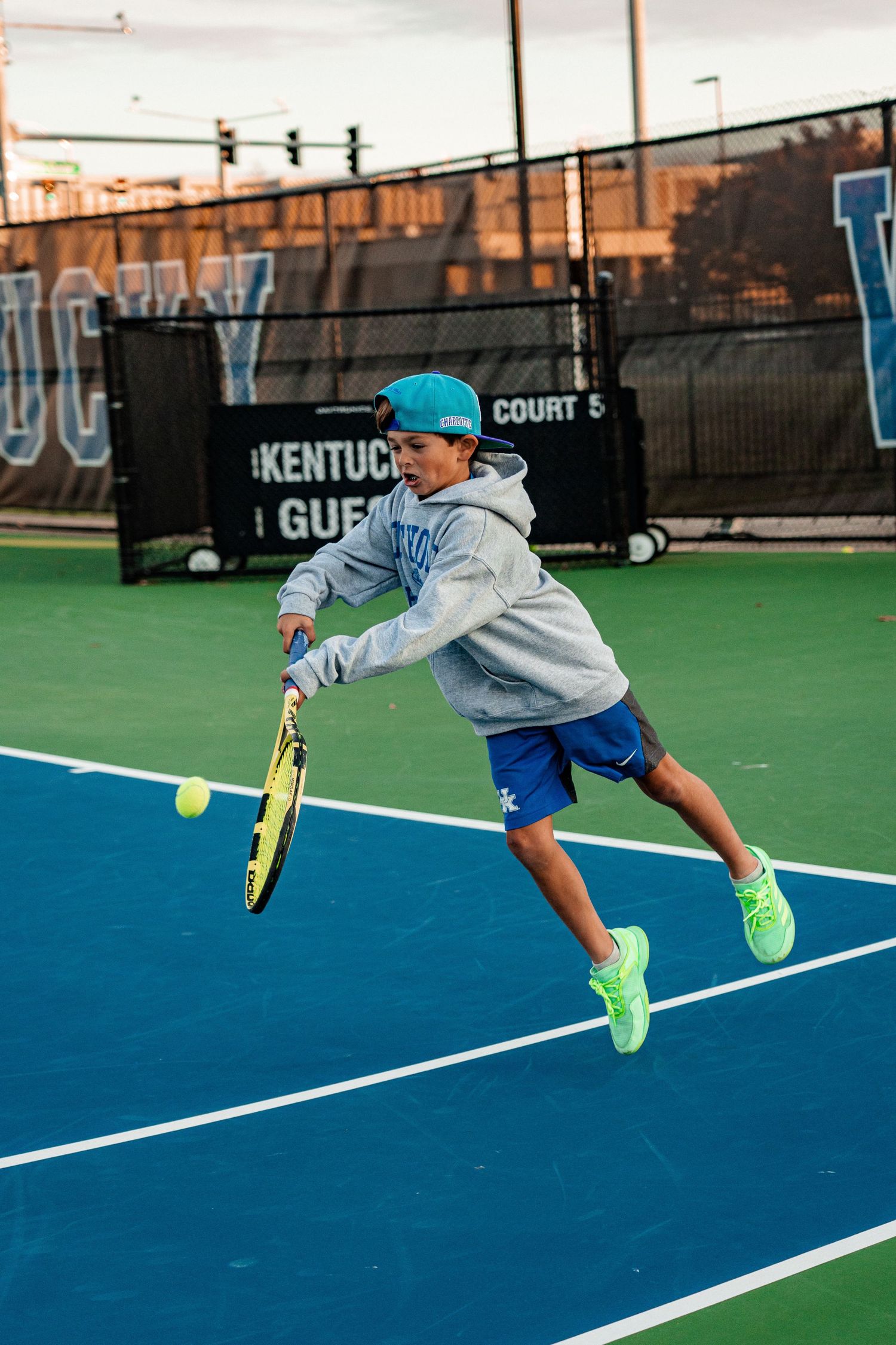 Tennis player in gray hoodie hits forehand shot while jumping on blue court.