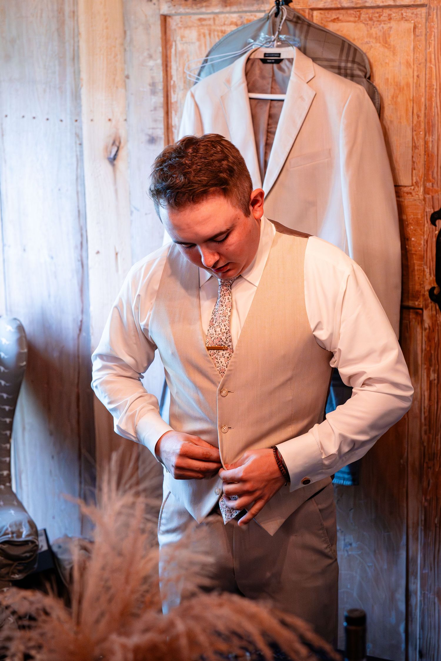 A groom adjusts his tie and vest while getting ready in a rustic wooden room.